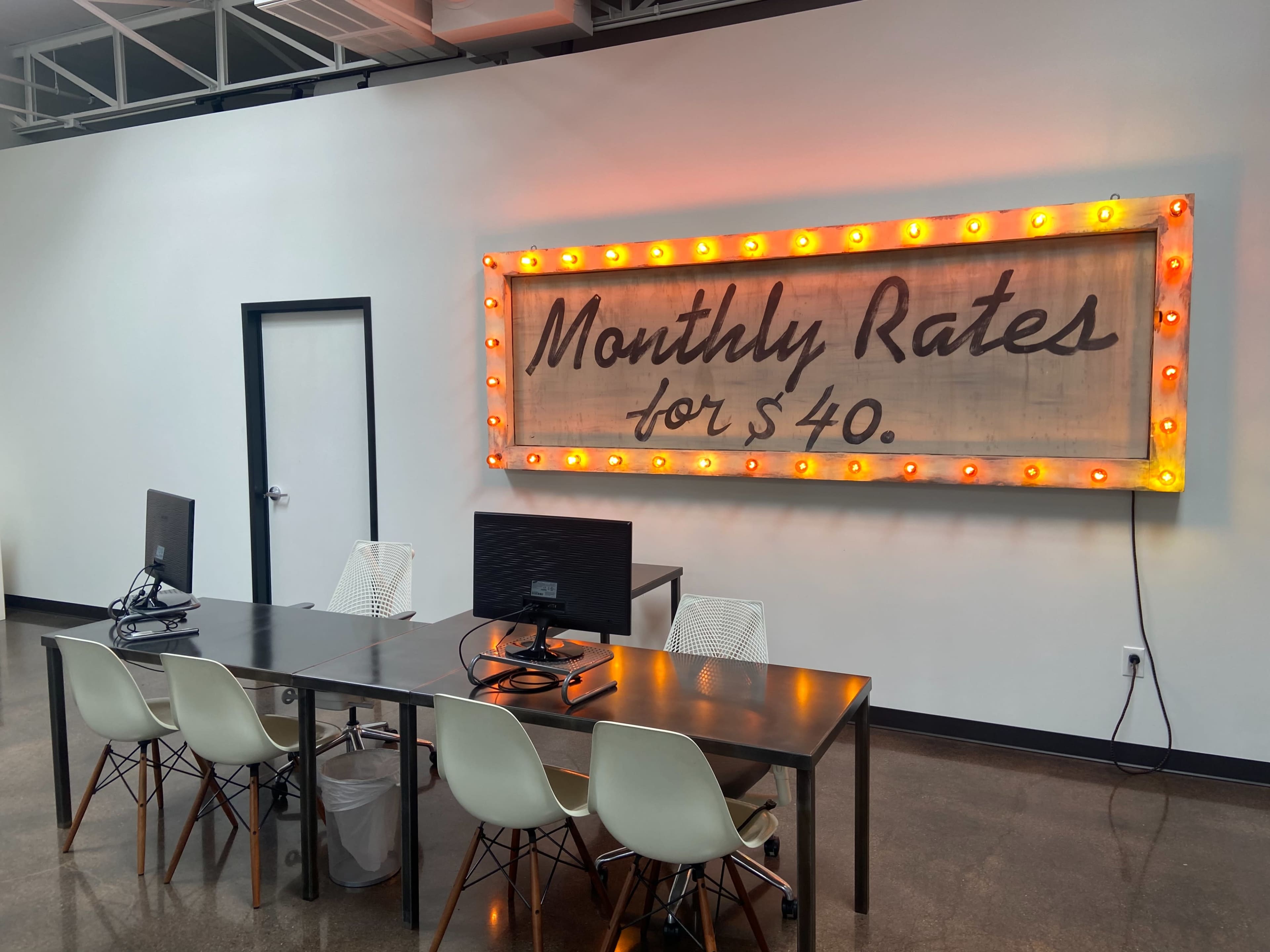 A modern workspace featuring a table with two computers and several white chairs, alongside a lit marquee sign that reads "Monthly Rates for $40."
