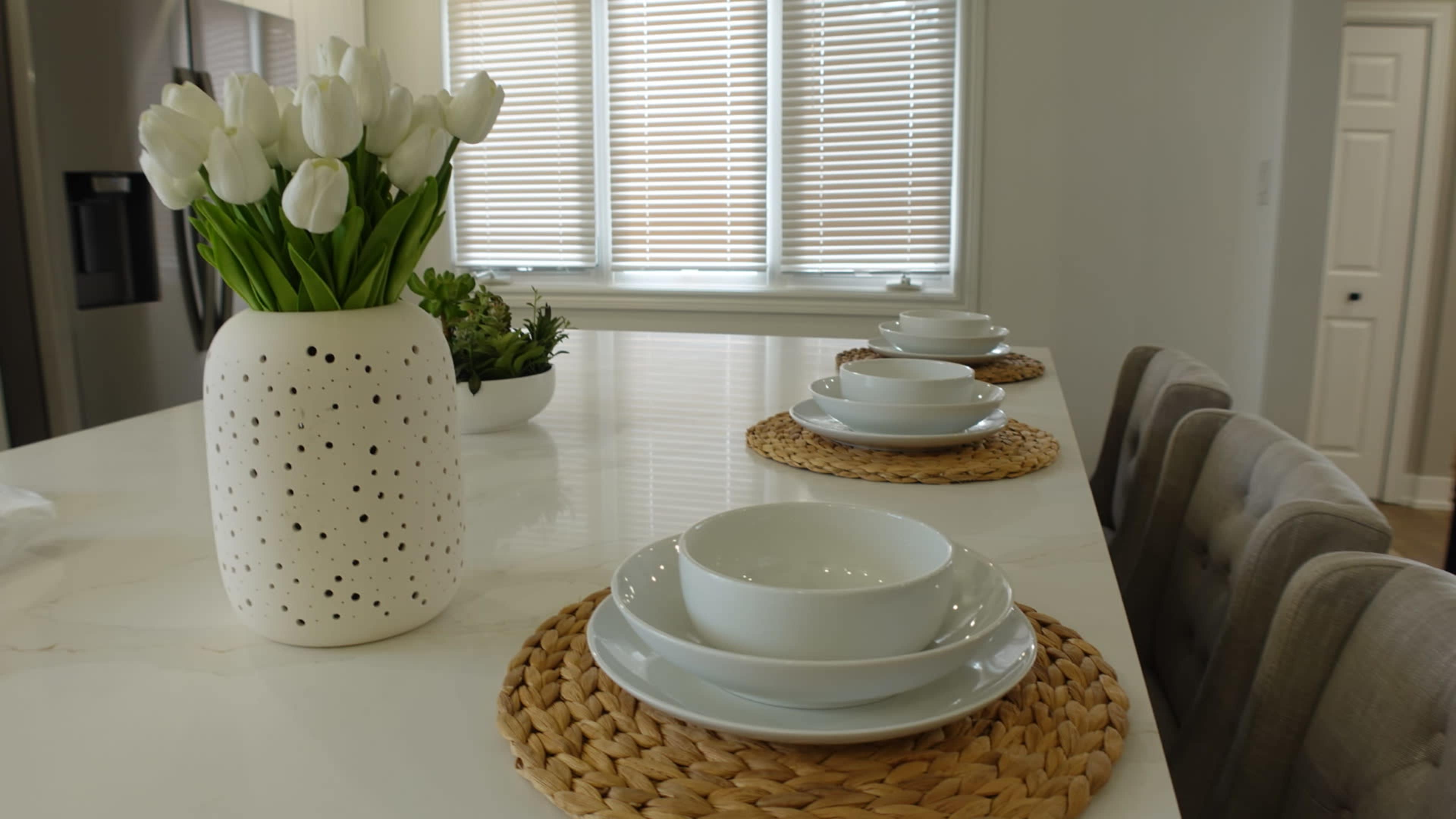 A dining area features a table set with white bowls on woven placemats, accompanied by a vase of tulips and a small plant in the background.