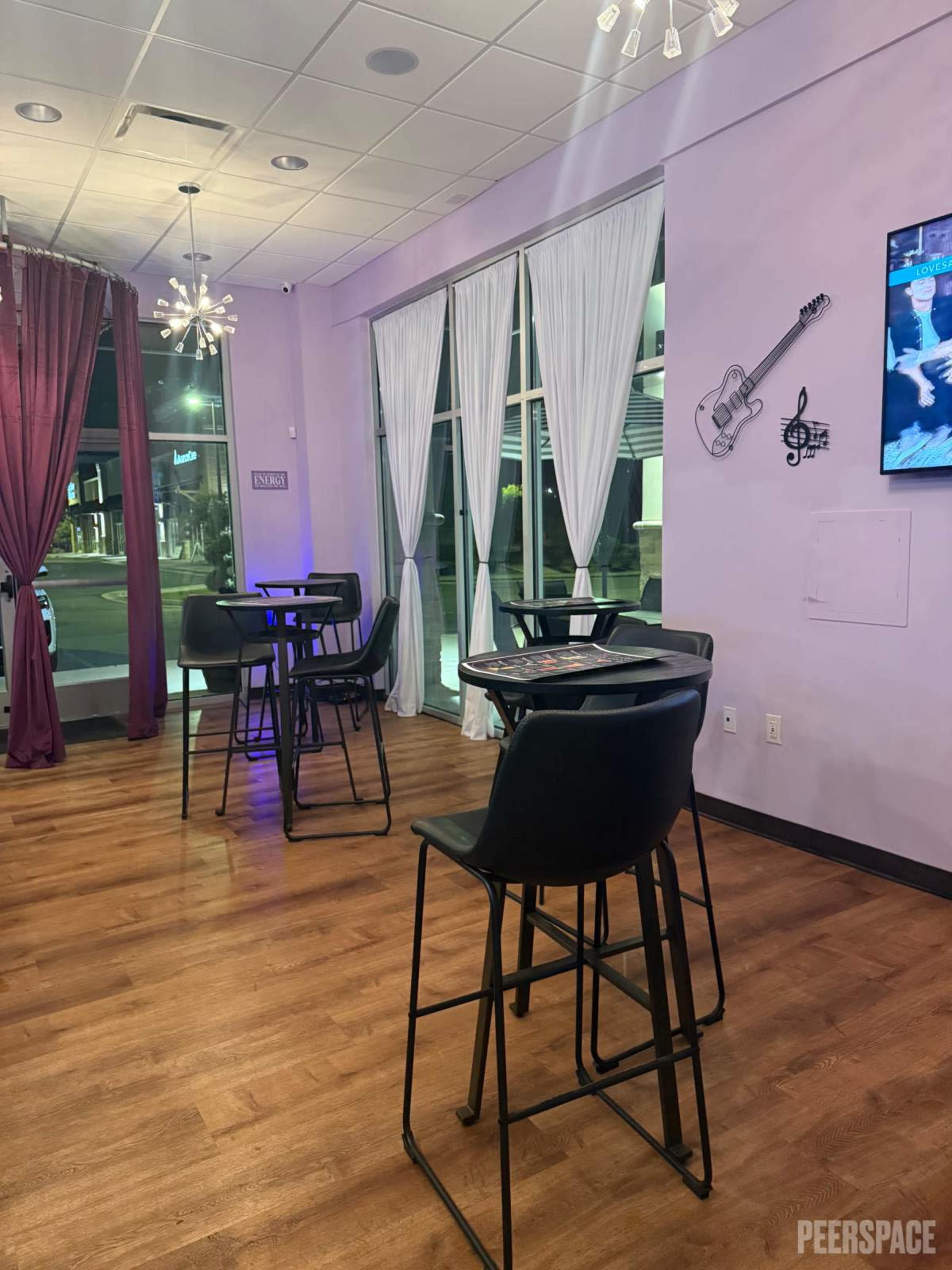 The image shows a brightly lit interior space with purple walls, black bar stools around high tables, and large windows covered with white and purple drapes.