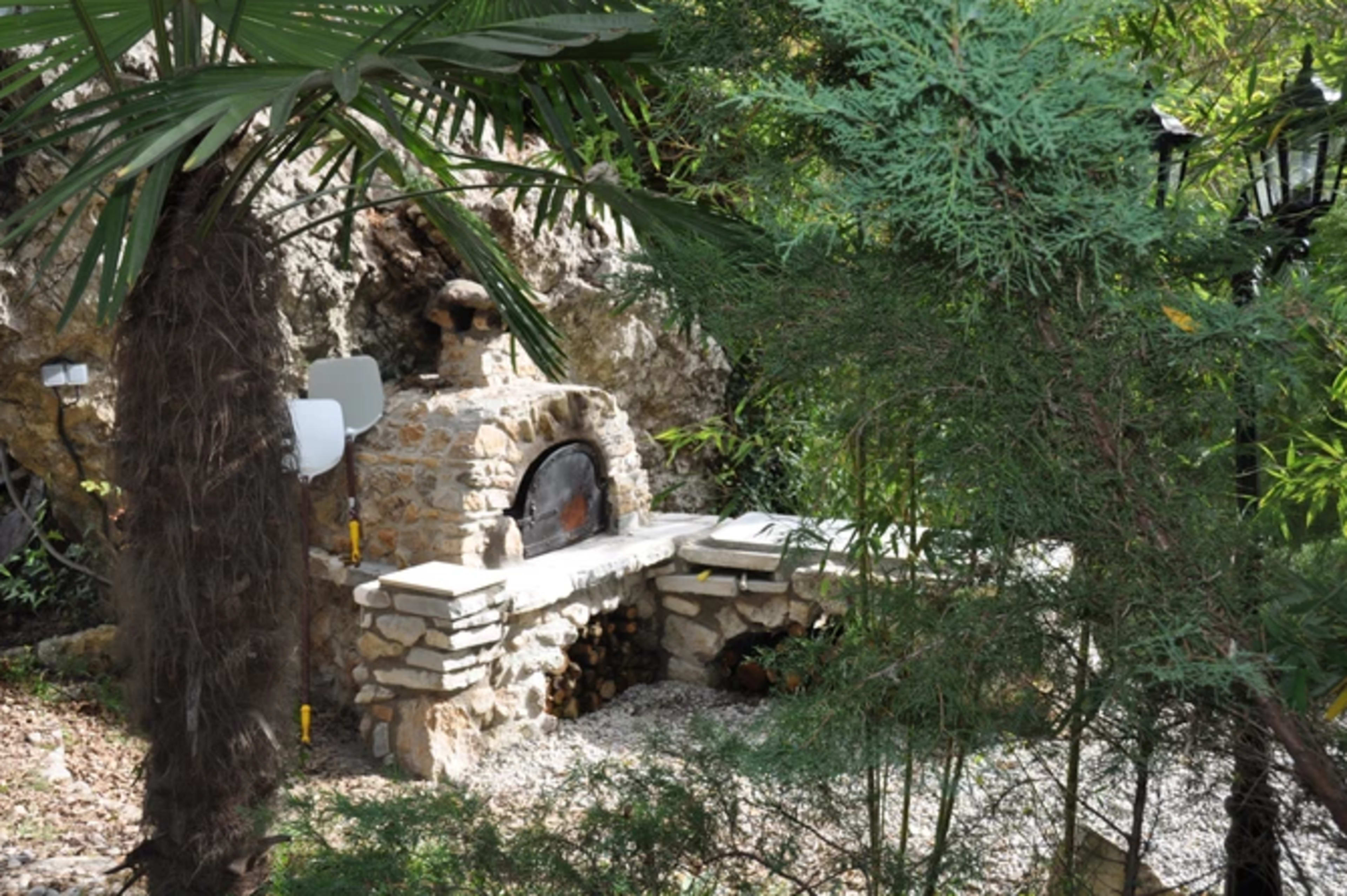 The image shows a stone outdoor oven nestled among greenery and surrounded by various plants.