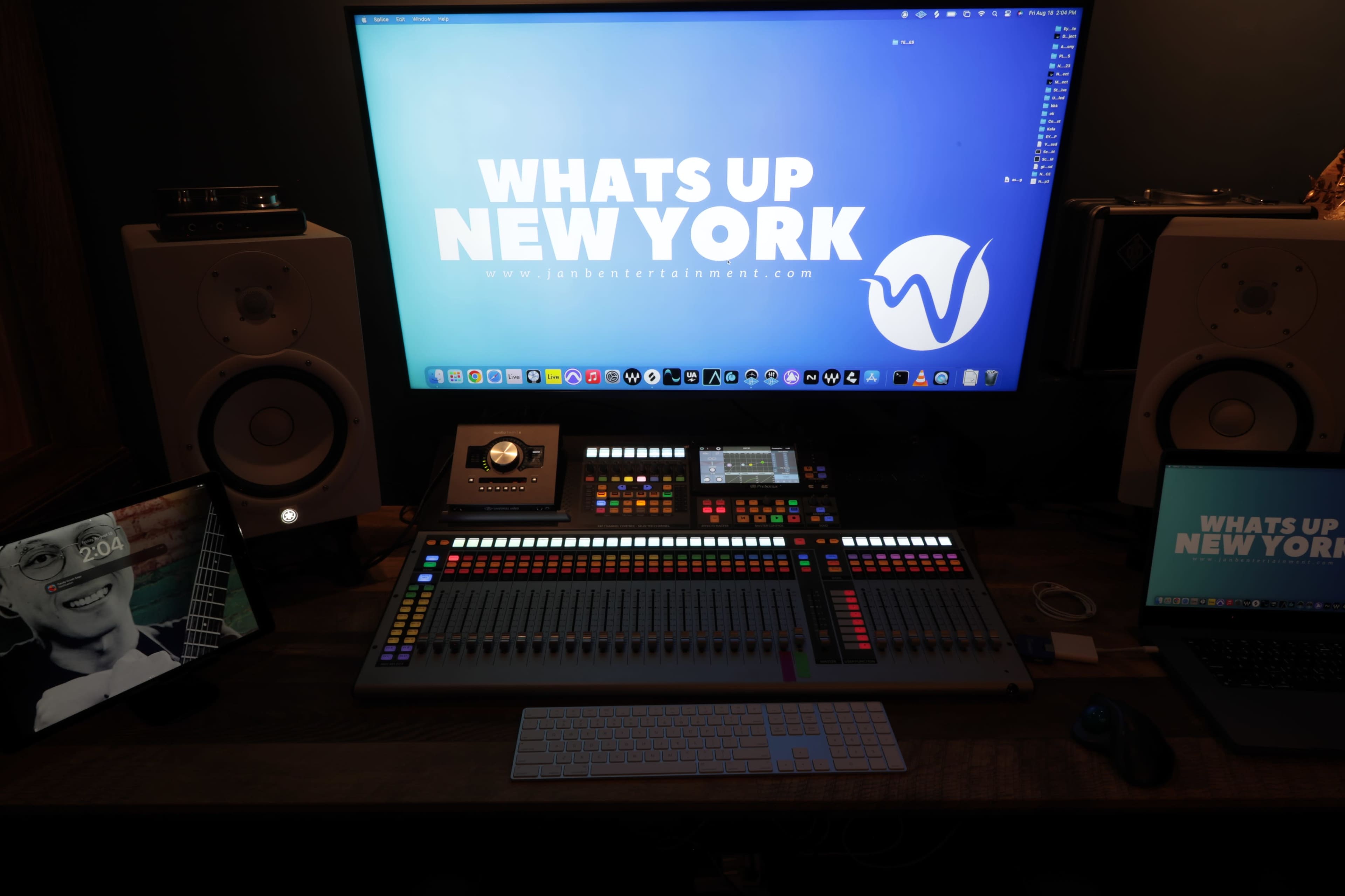 A music production workstation featuring a large monitor, audio speakers, a digital mixing console, and two laptops with a "What's Up New York" graphic.