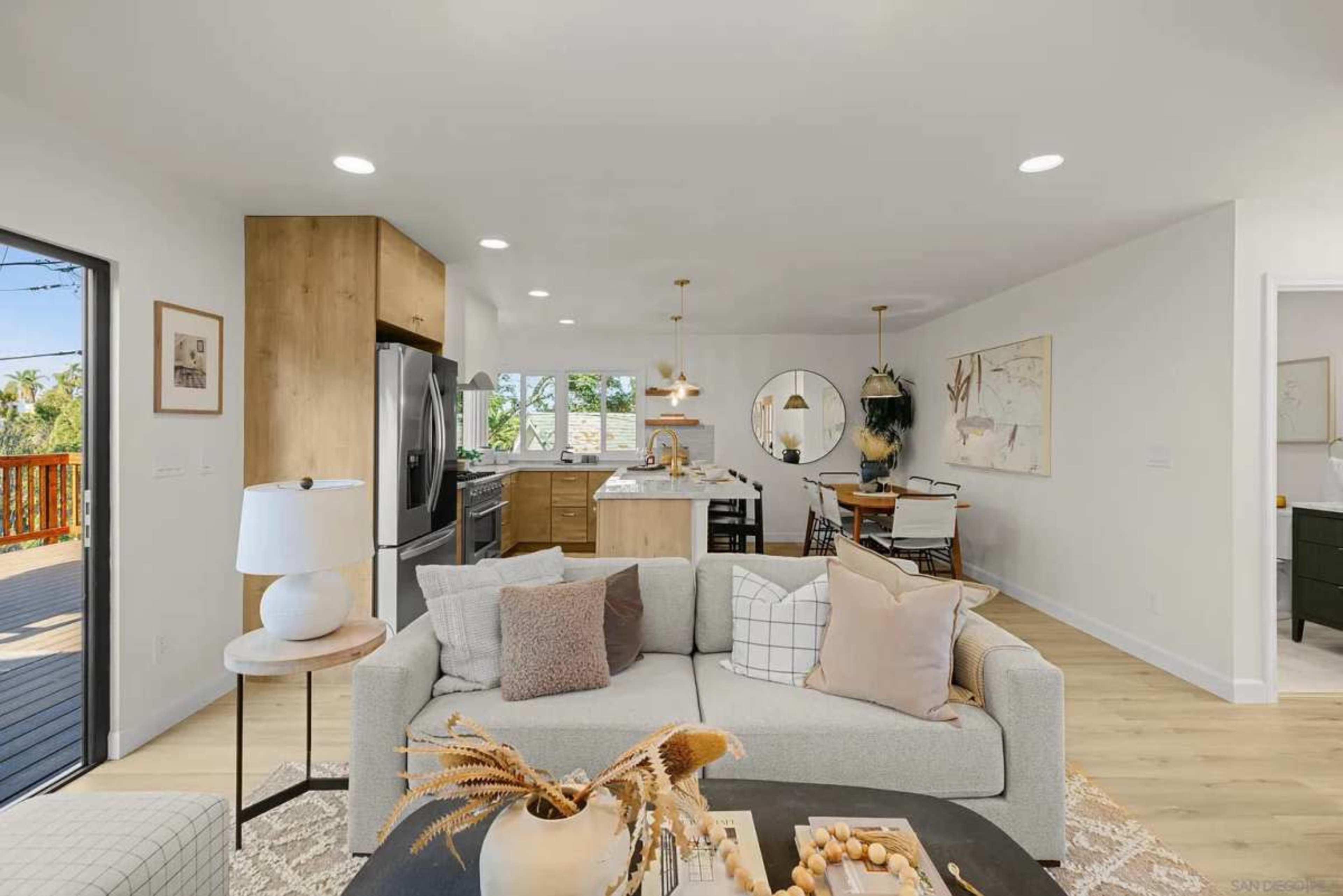 The image shows a modern open-concept living space with a light-colored sofa, a dining area, and a kitchen featuring wood cabinetry and stainless steel appliances.