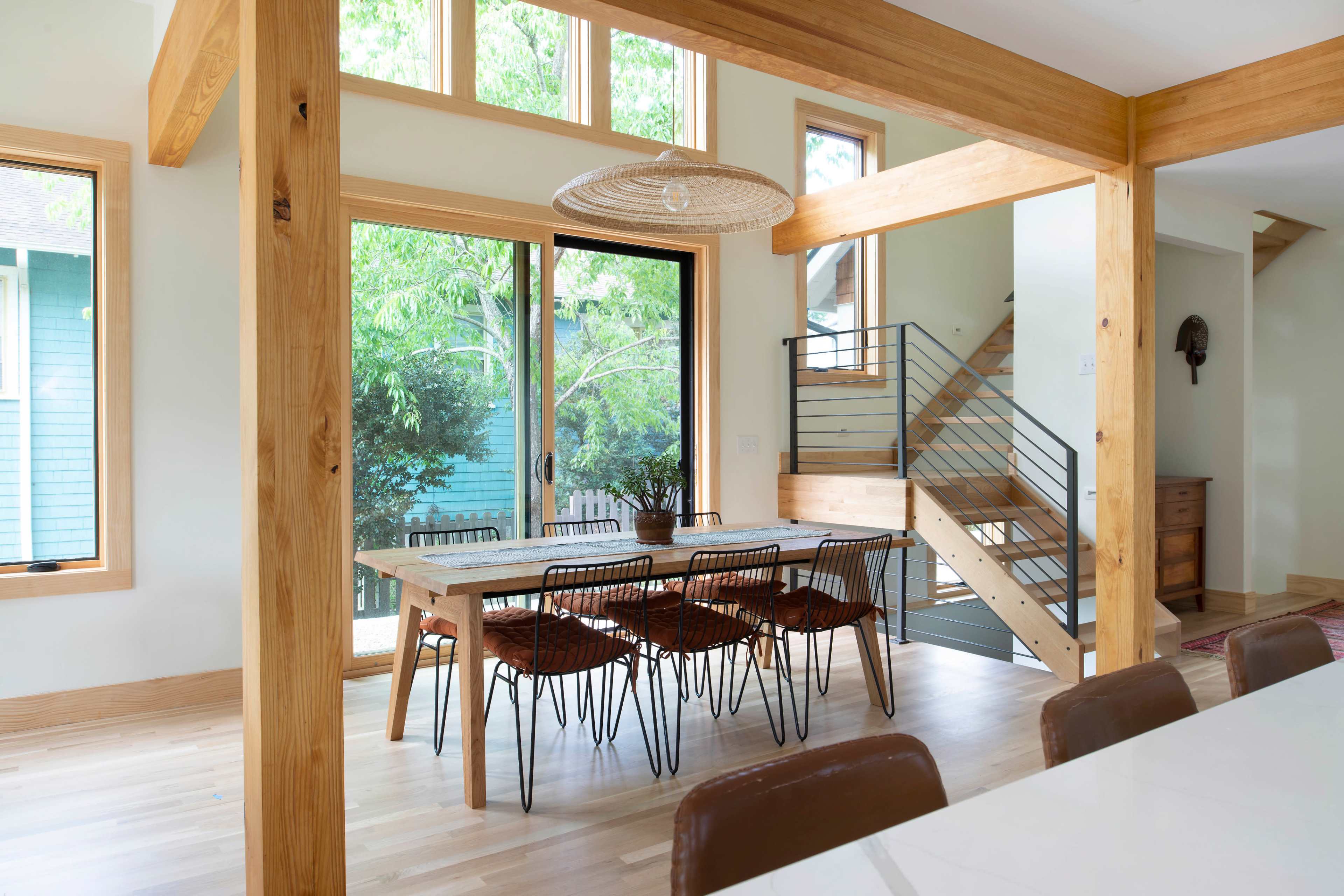 A dining area features a wooden table surrounded by black wire chairs, with large windows allowing natural light and views of greenery.