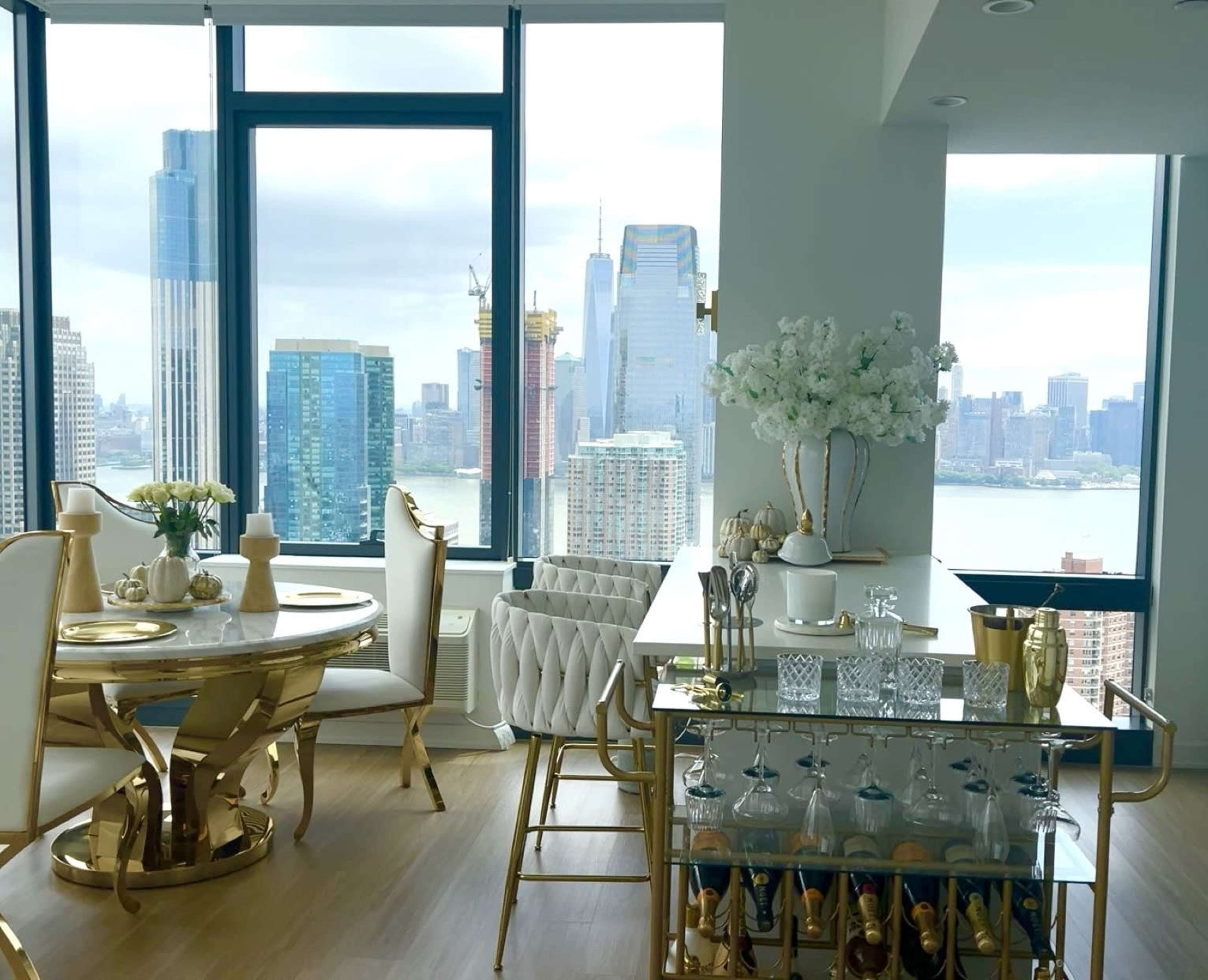 A modern dining area features a round table with elegant seating and a bar cart, overlooking a city skyline with tall buildings and water in the distance.