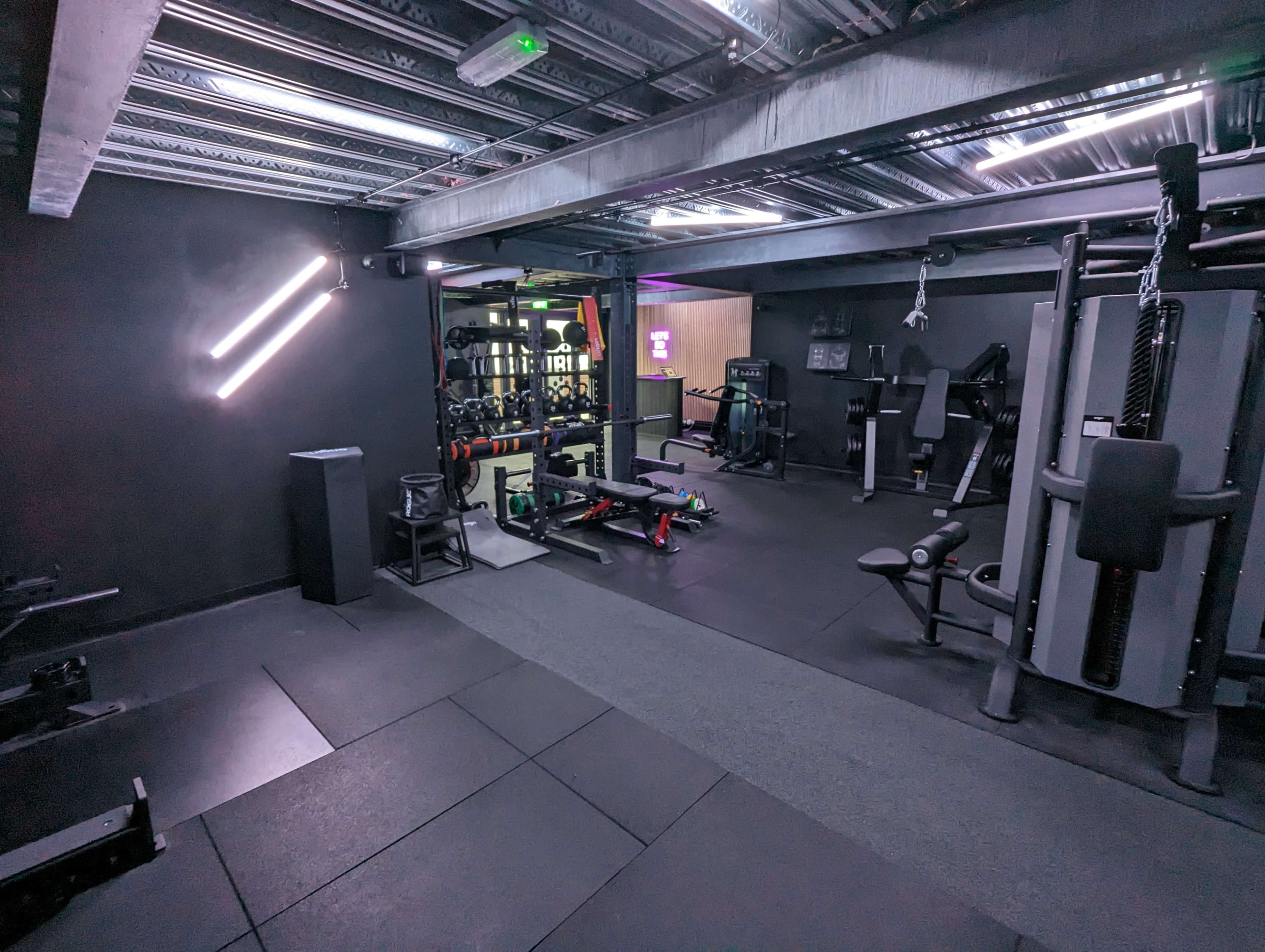 A modern gym interior with various fitness equipment arranged on a dark floor under artificial lighting.