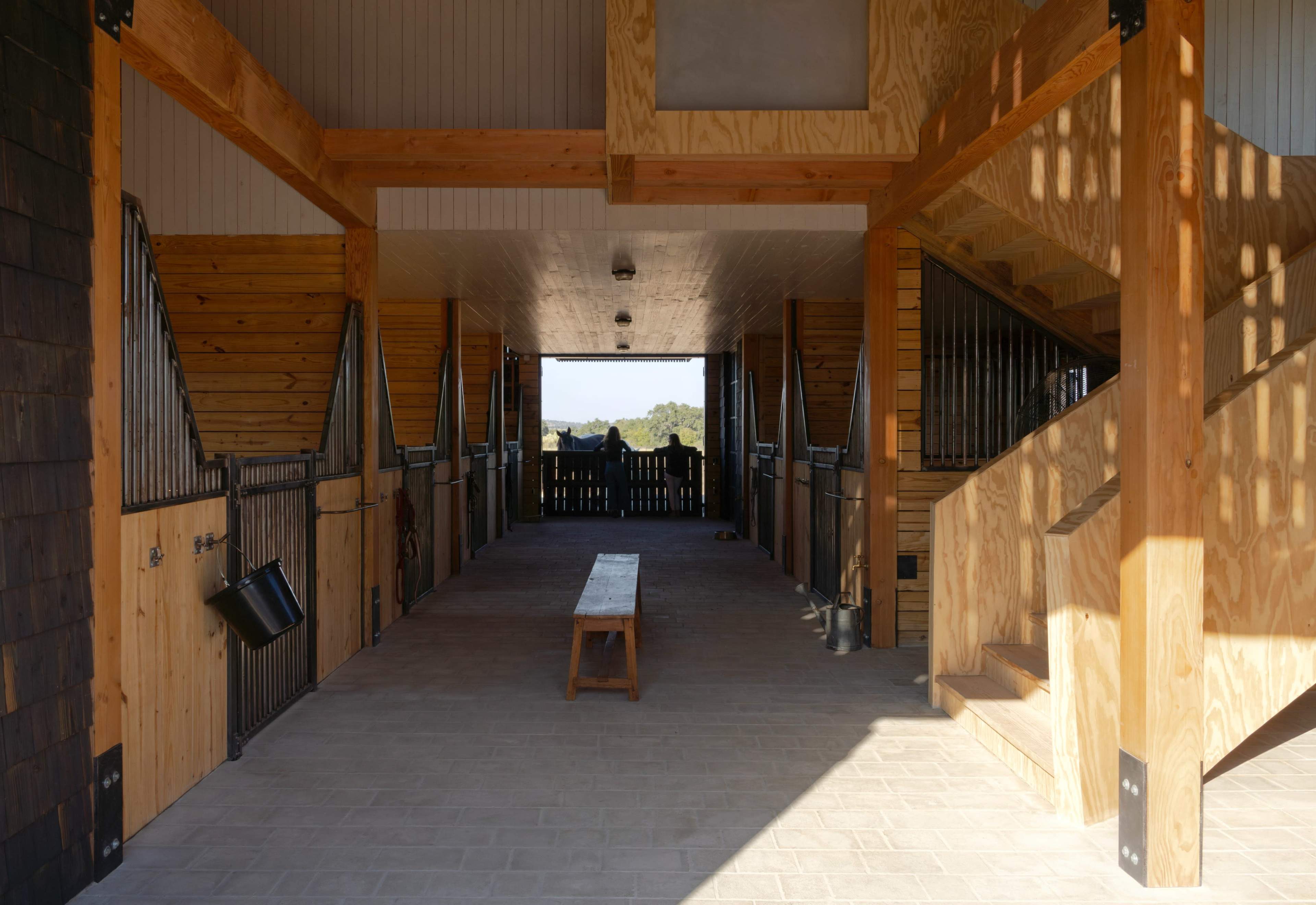 The image shows the interior of a barn with wood and metal features, including stalls on either side and a bench in the center.