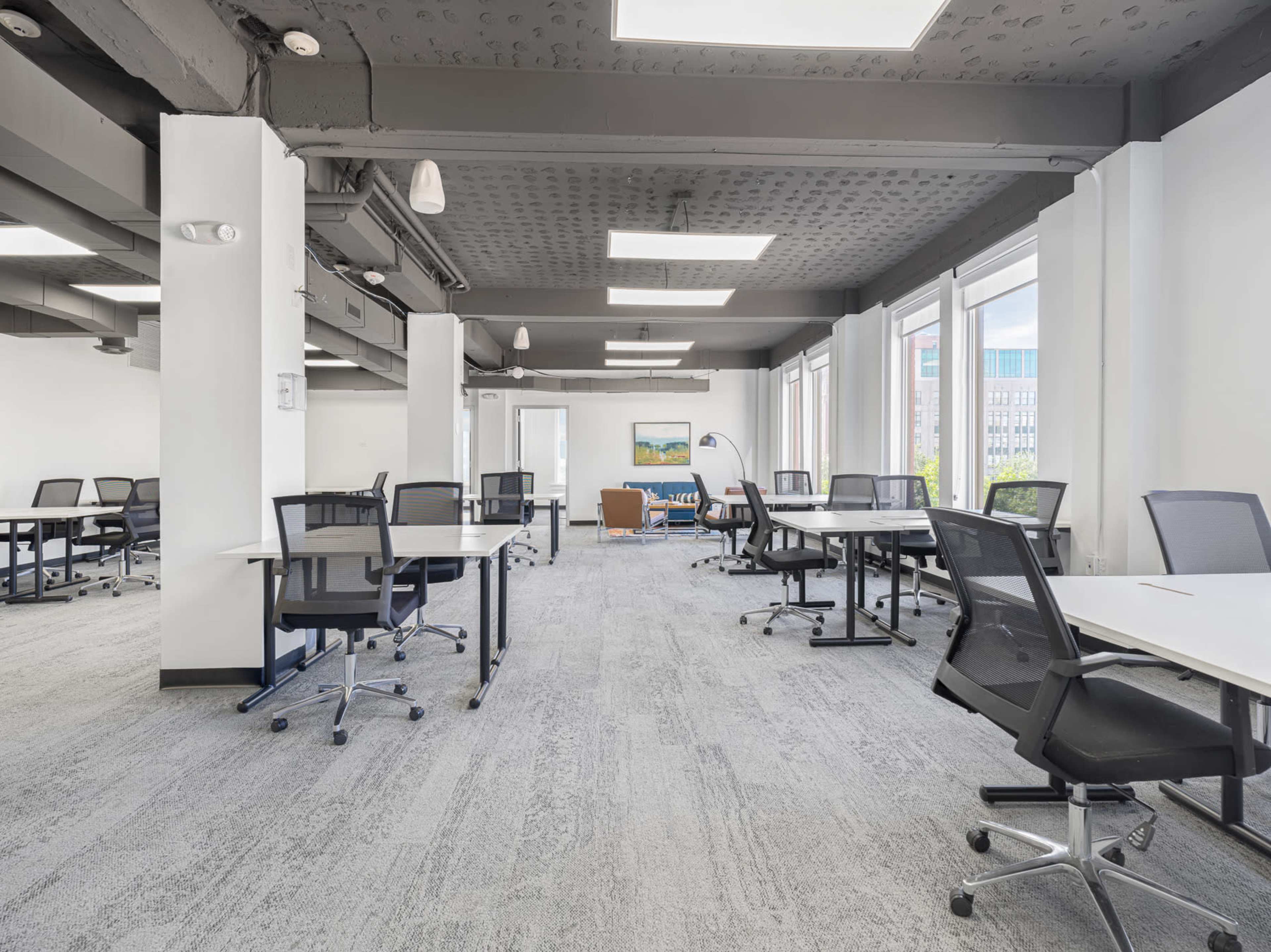 The image shows a modern office space with several empty desks arranged in a well-lit environment featuring large windows.