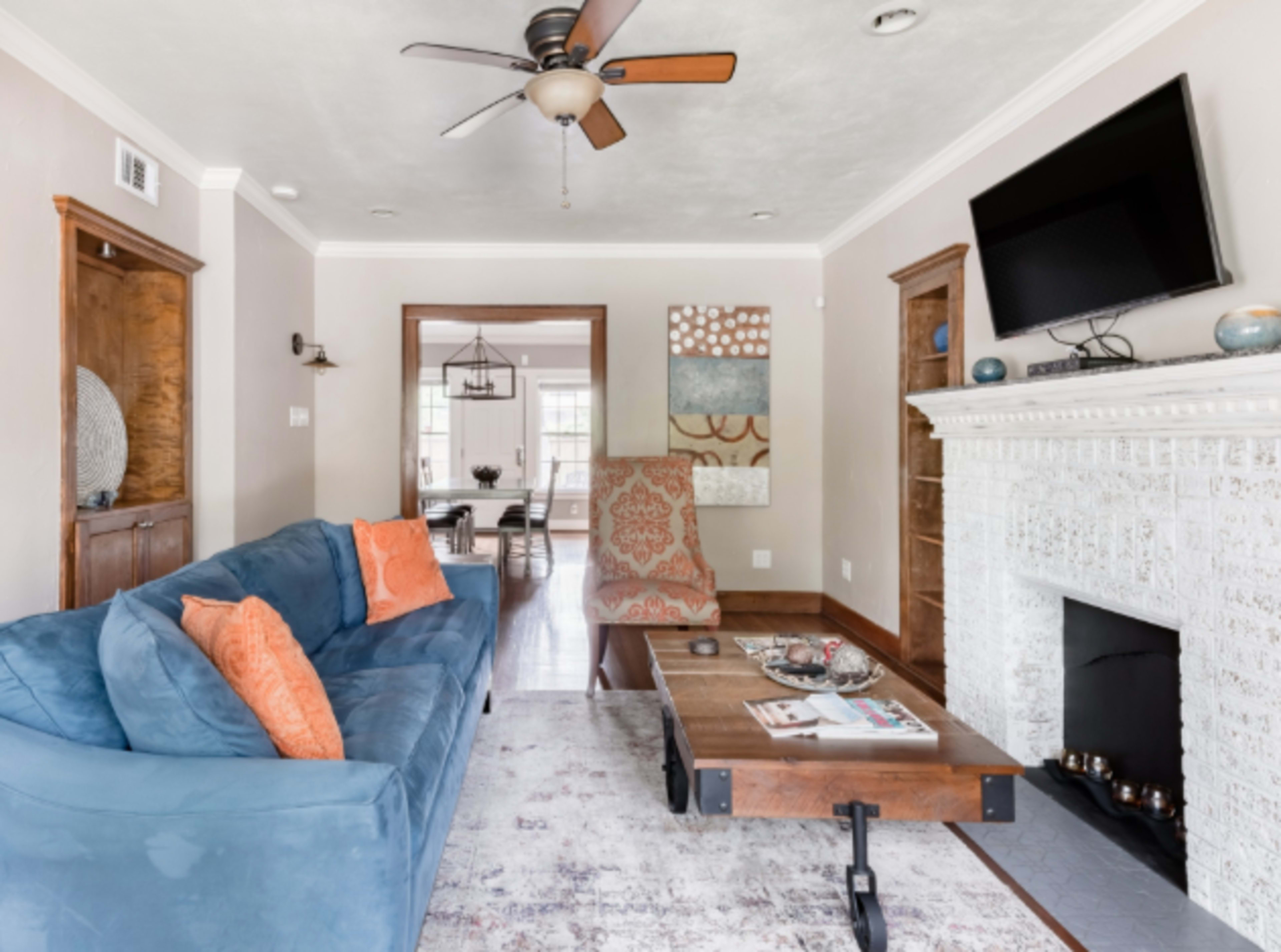 The living room features a blue sofa with orange pillows, a wooden coffee table, and a fireplace, with a view of a dining area in the background.