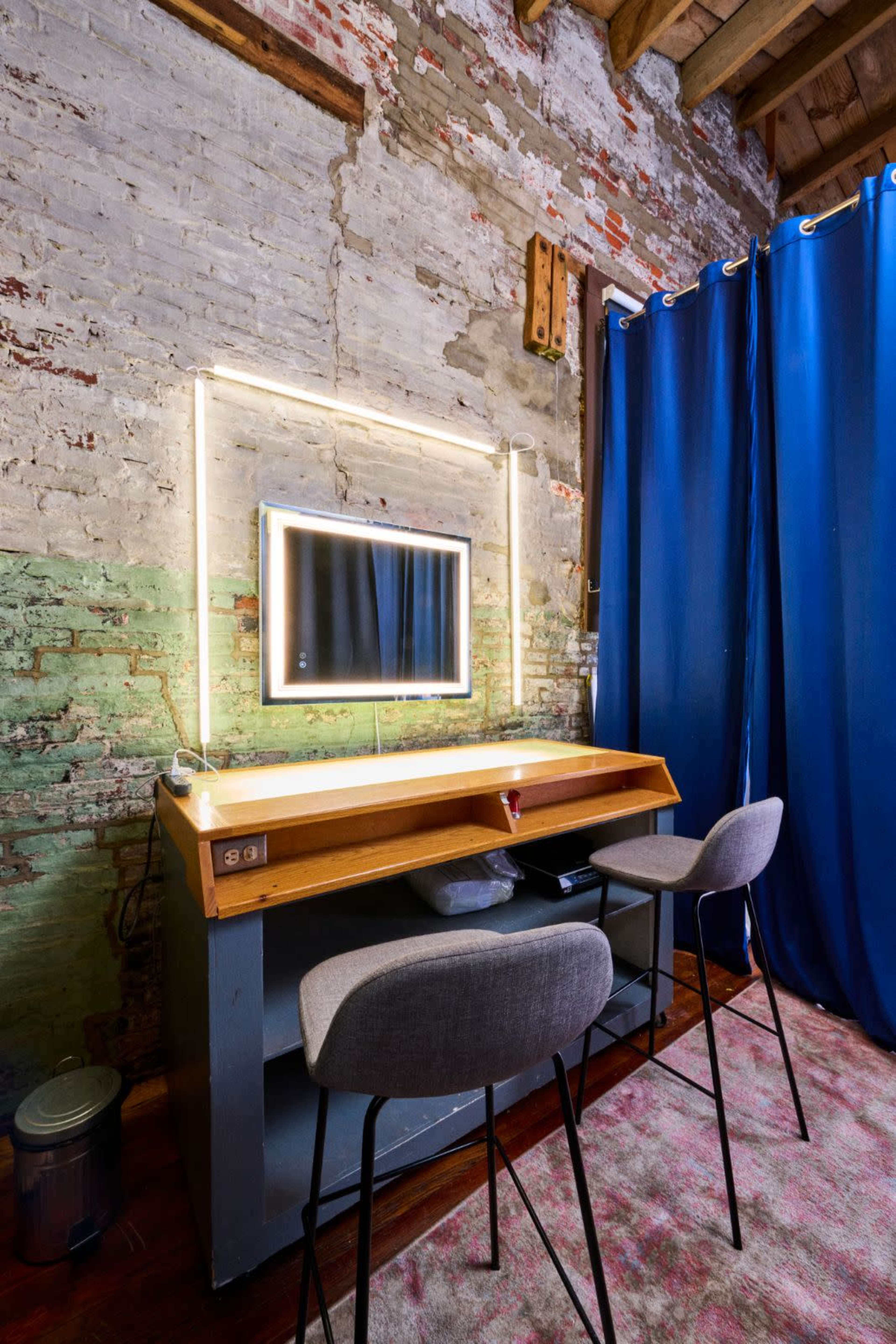 A wooden vanity with two gray chairs and a mirror is situated against a brick wall, illuminated by a light strip above it, with blue curtains on either side.