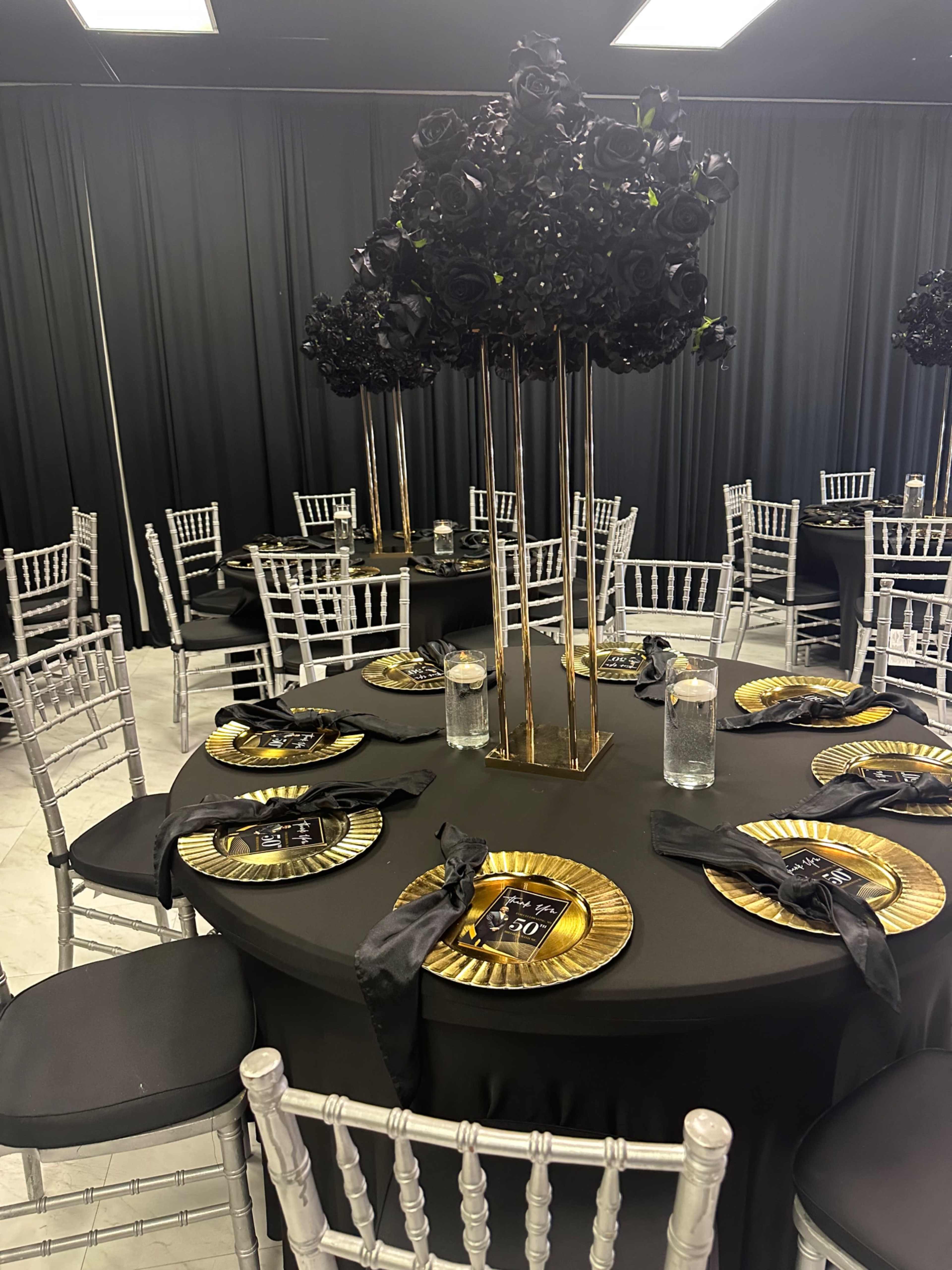 A dining area is set with round black tables decorated with gold and black plates, floral centerpieces, and silver chiavari chairs against a black curtain backdrop.