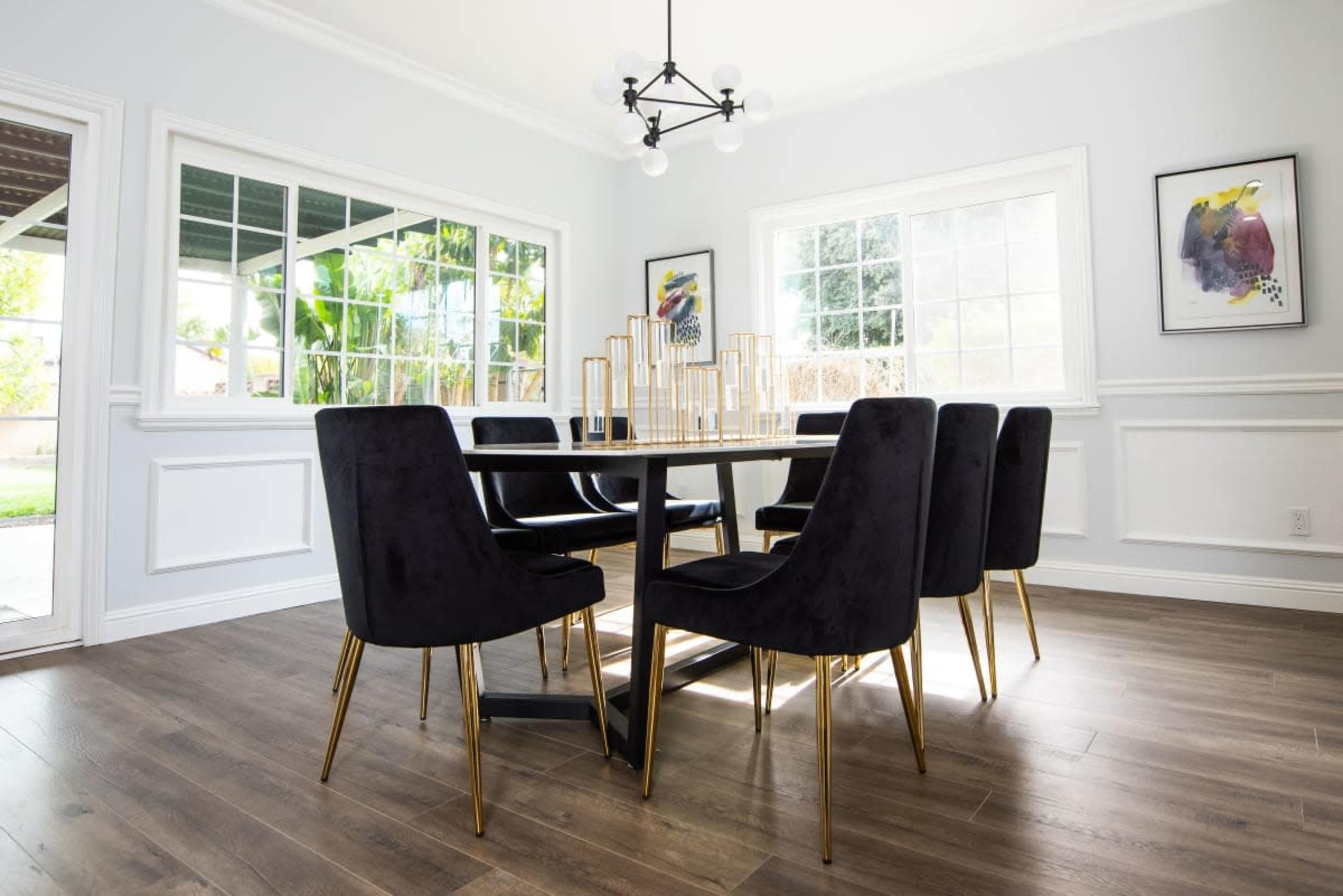 A dining room features a rectangular table surrounded by six black chairs with gold legs, set under a modern light fixture and illuminated by natural light from large windows.