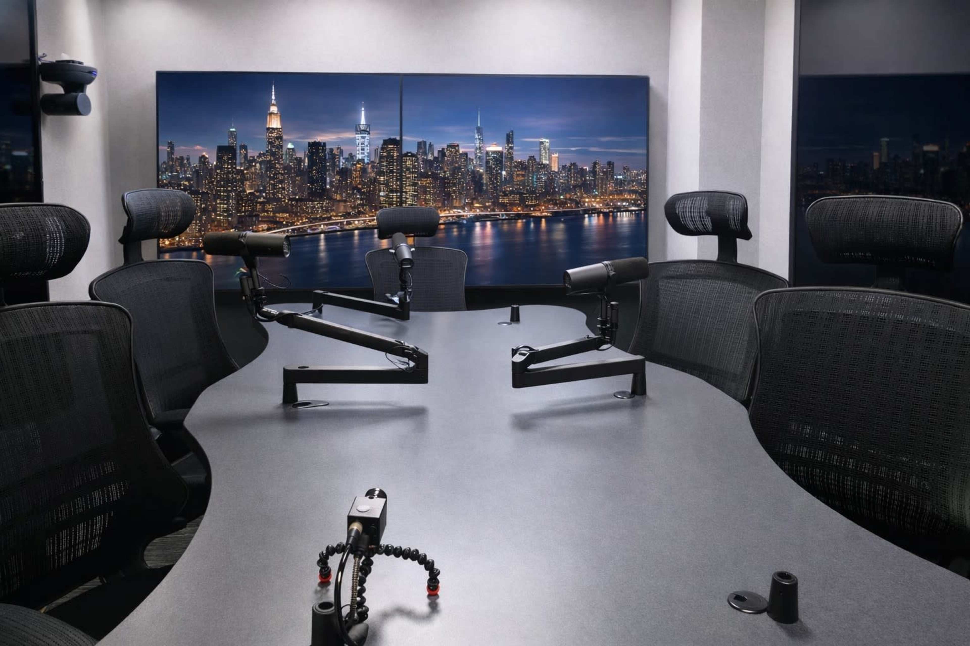 A modern conference room features a sleek oval table surrounded by ergonomic chairs and two camera mounts facing a city skyline backdrop.