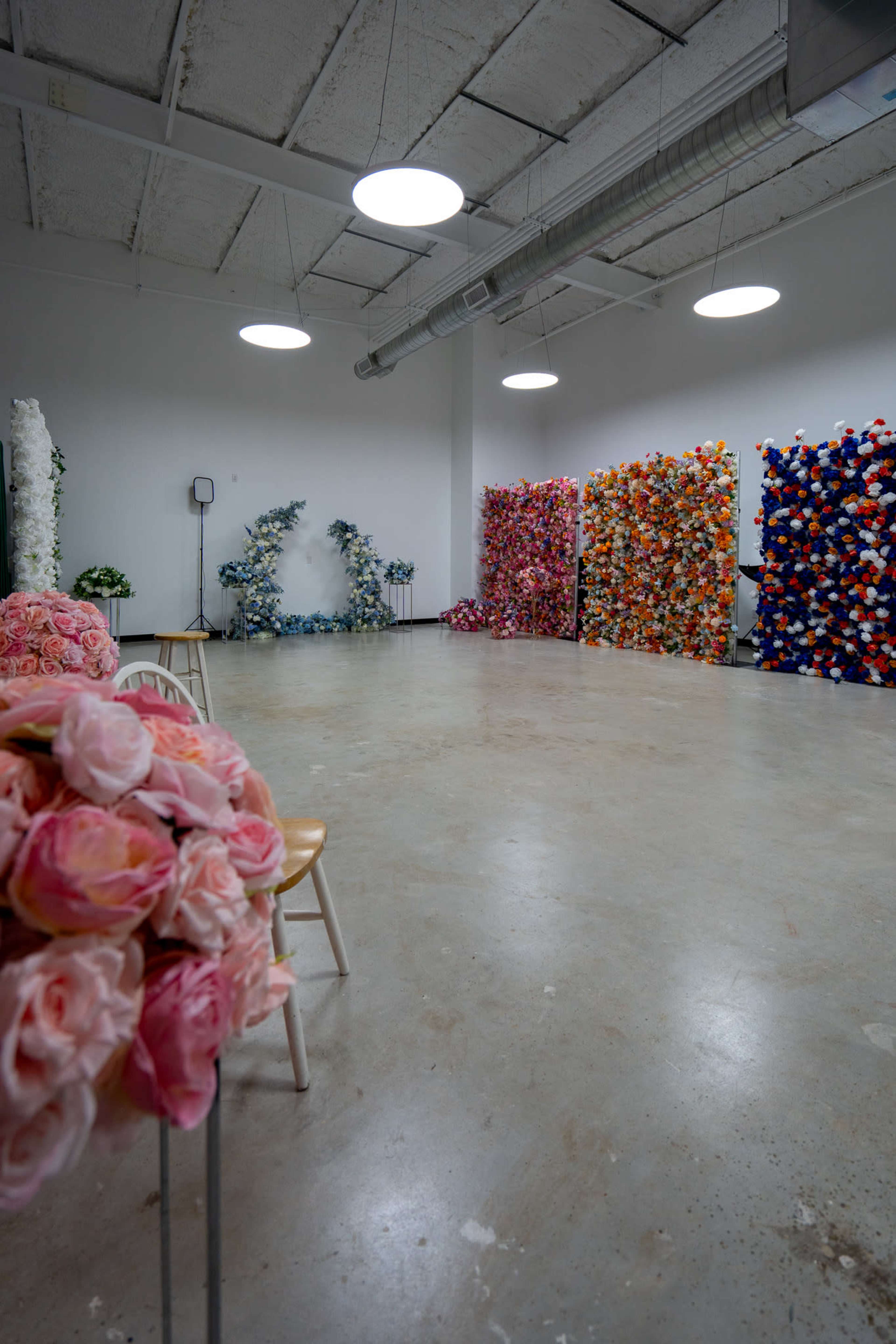 The image shows a spacious, well-lit room with multiple walls covered in artificial floral arrangements, while a few wooden chairs are positioned in the foreground.