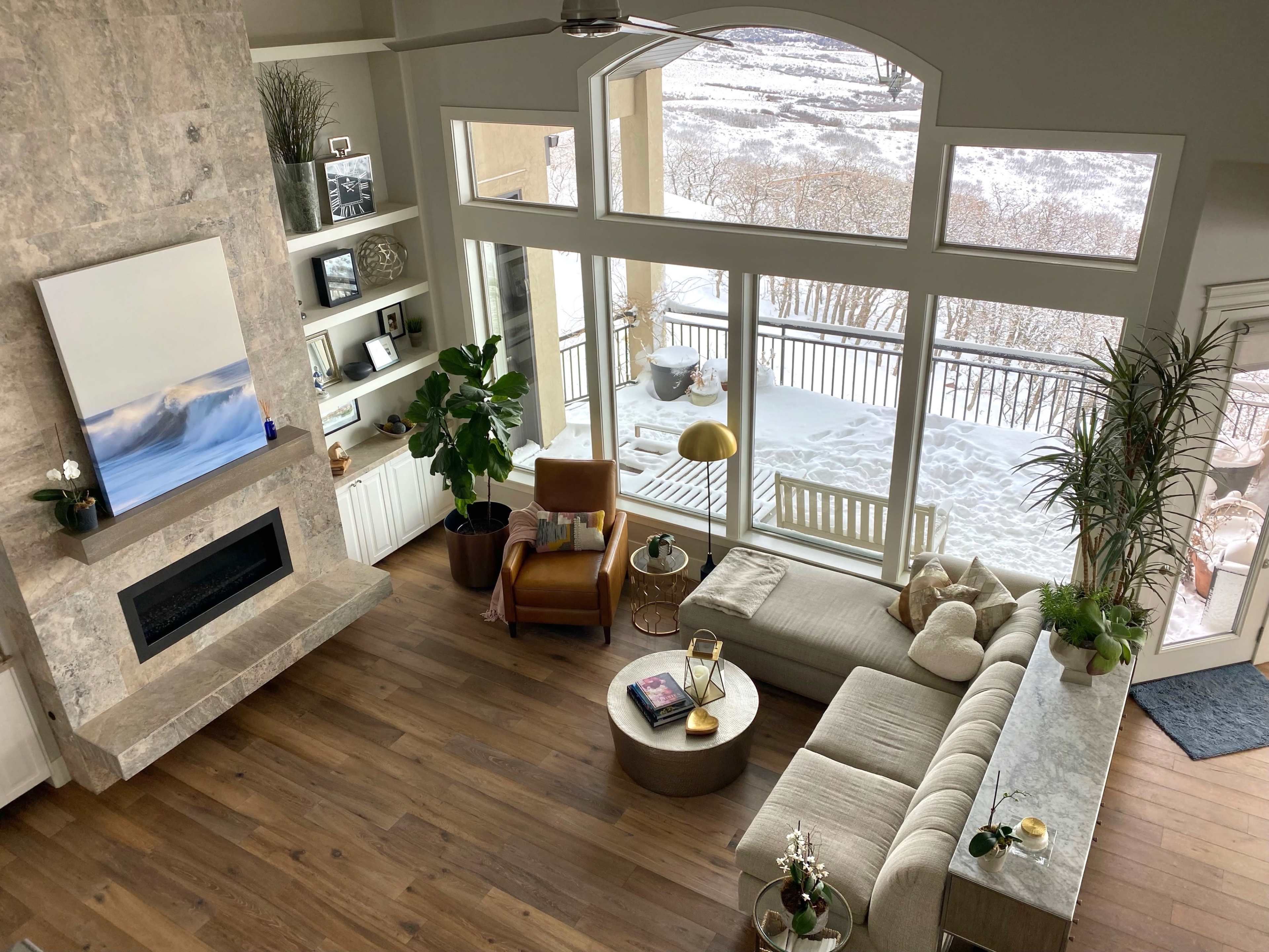 A spacious living room with a large window overlooking a snowy landscape, featuring a modern fireplace, a beige sectional sofa, a wooden accent chair, and decorative plants.
