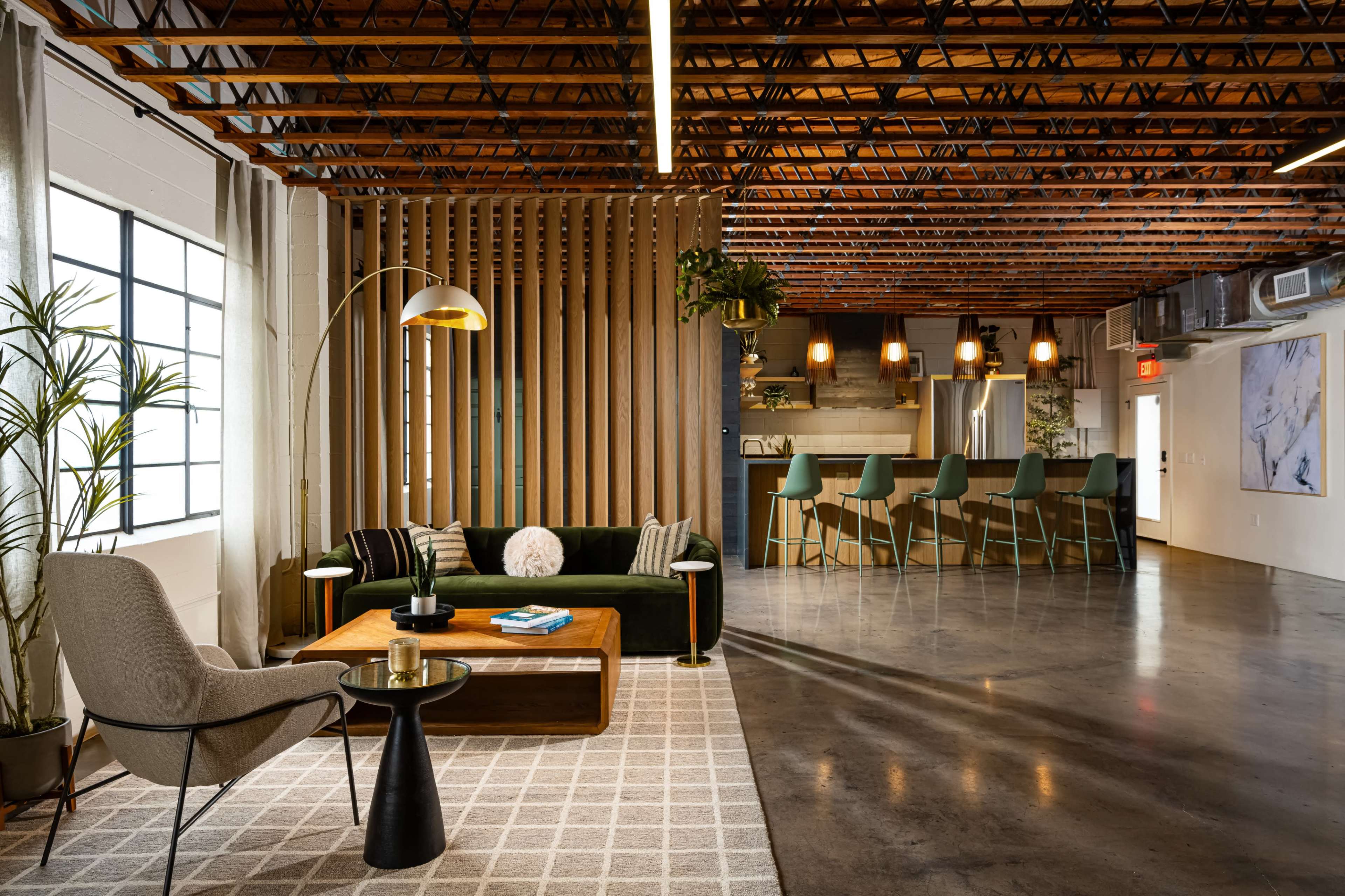 The image shows a modern interior space featuring a green couch, an armchair, a wooden coffee table, and a kitchen area with barstools under industrial-style lighting.