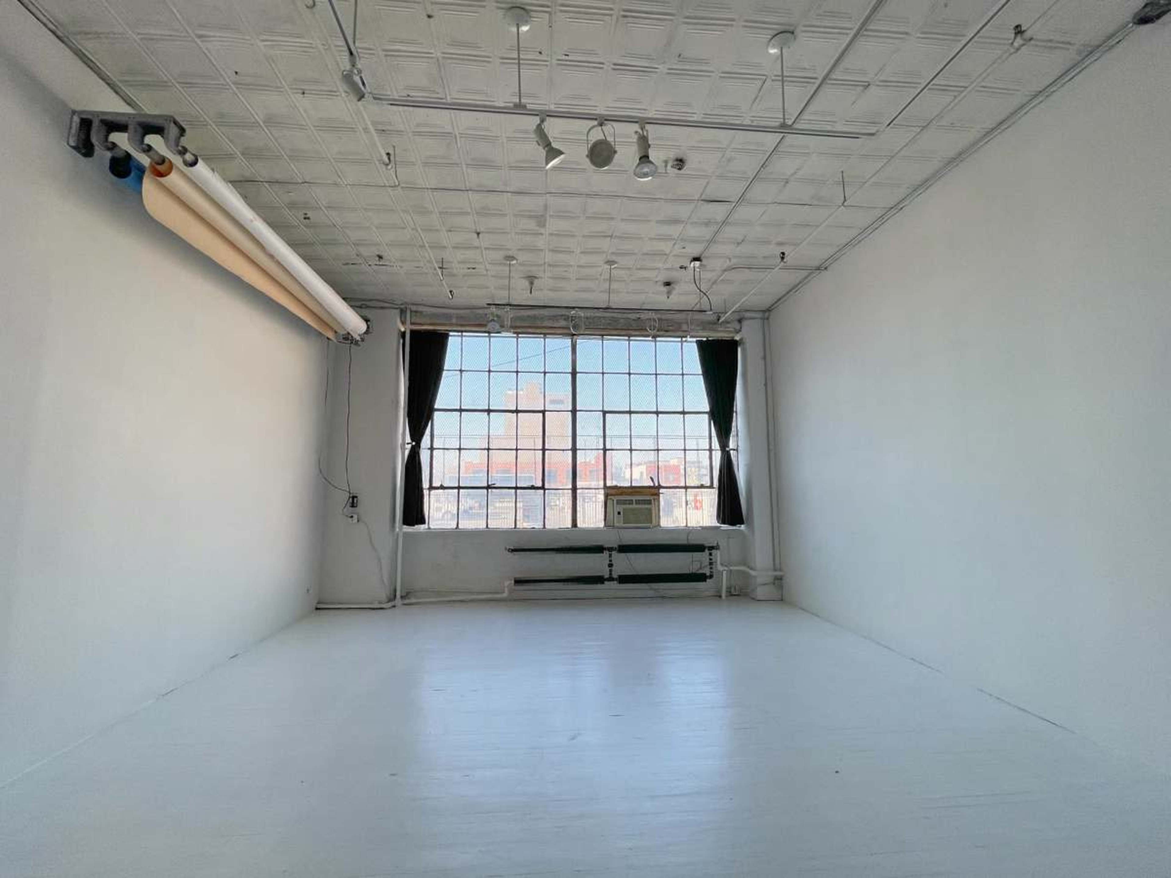 The image shows an empty, bright studio space with white walls, a large window, and an air conditioning unit beneath the window.