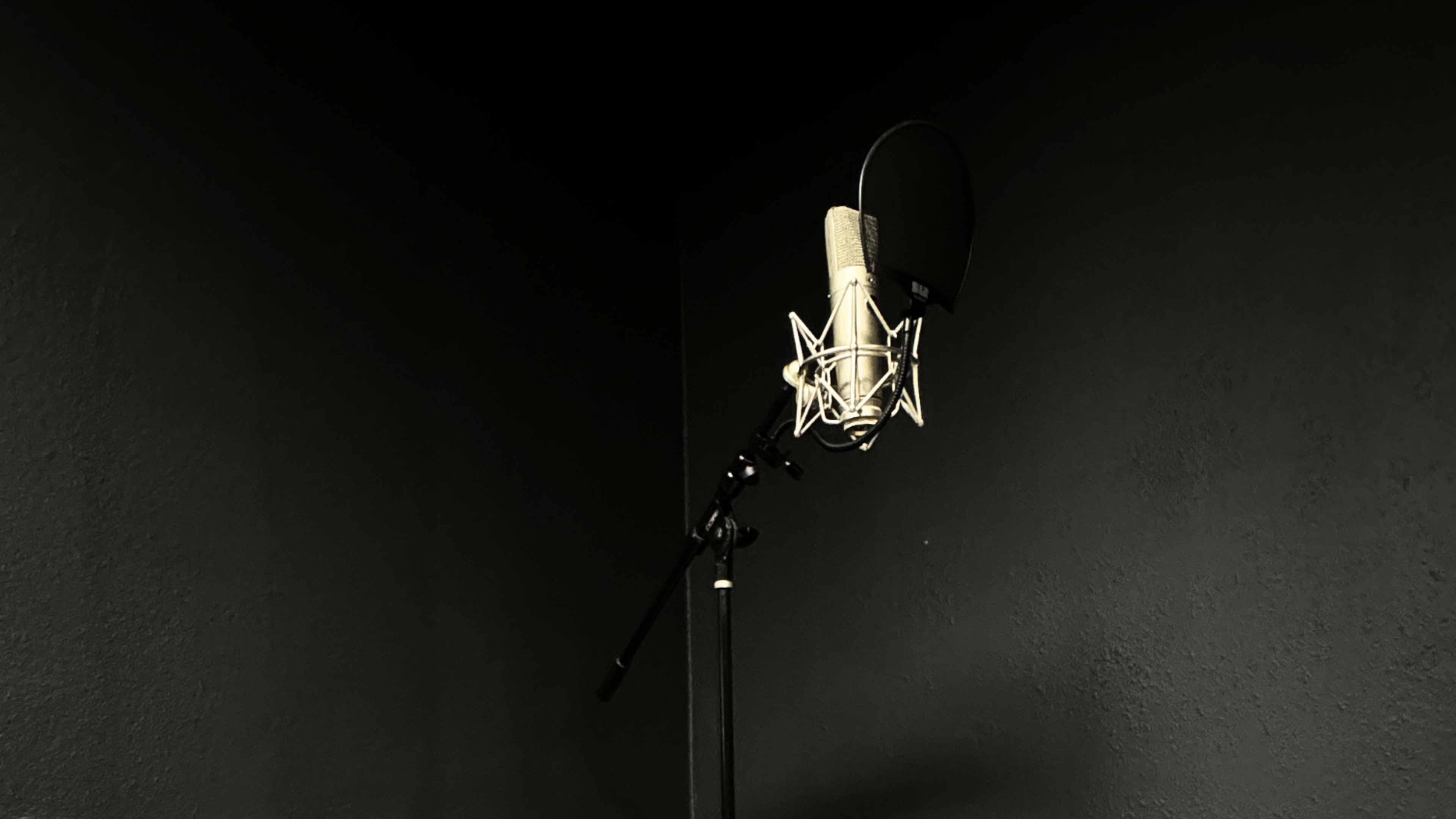 A microphone stands on a black stand in a dimly lit corner against a dark wall.