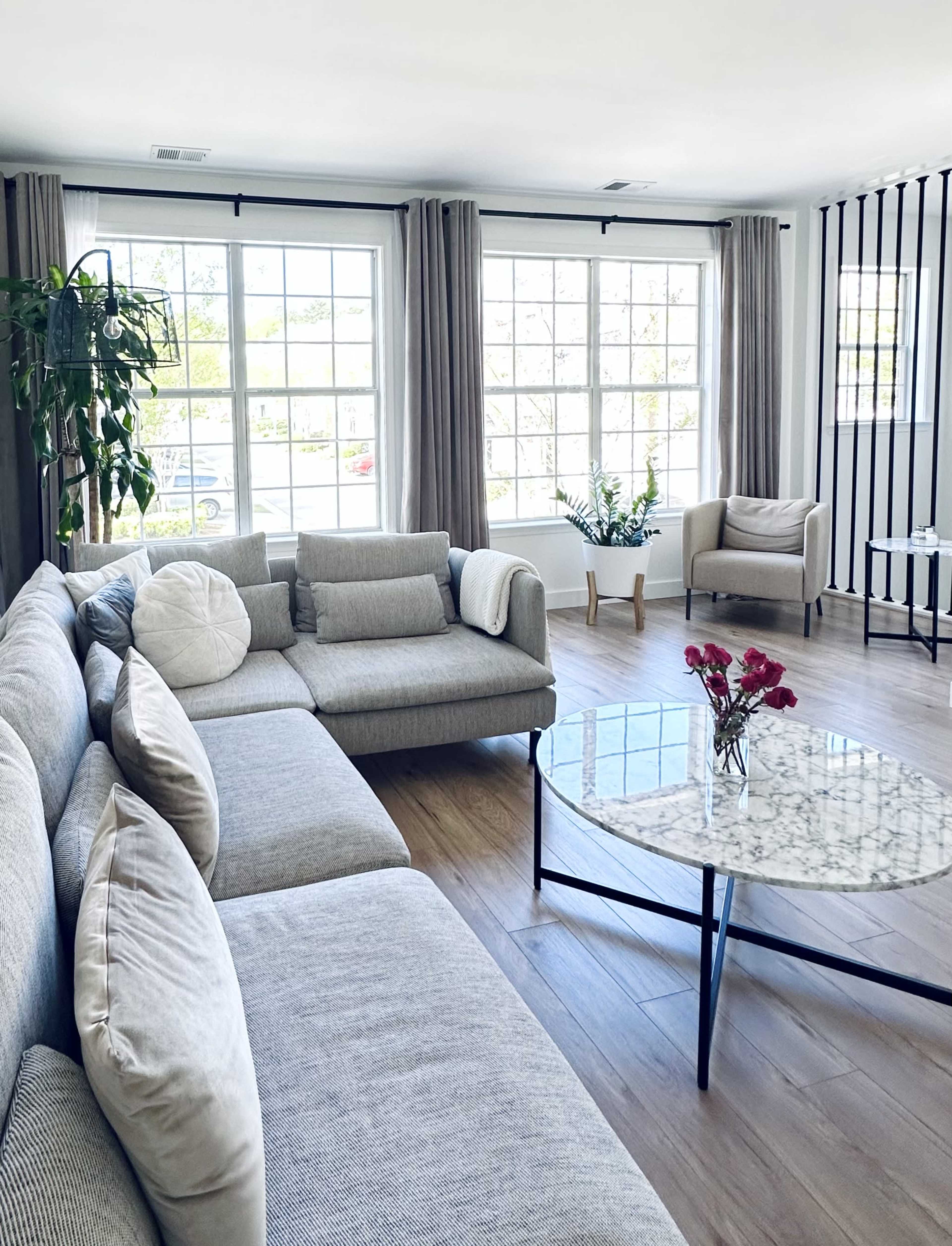 A bright living room with a large sectional sofa, a marble coffee table with flowers, and windows that let in natural light.