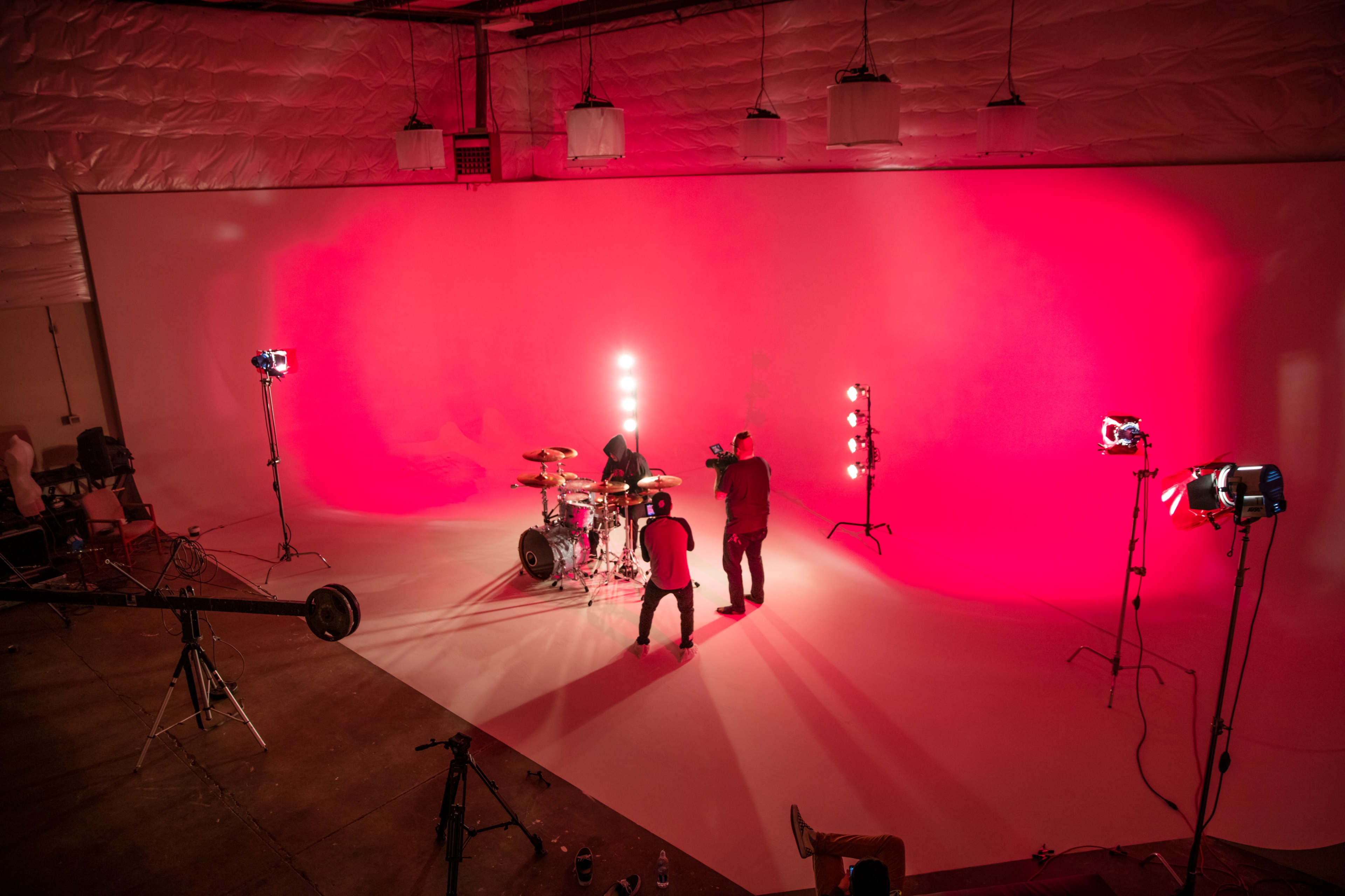A band performs on a stage with a bright pink backdrop and various lighting equipment set up around them.