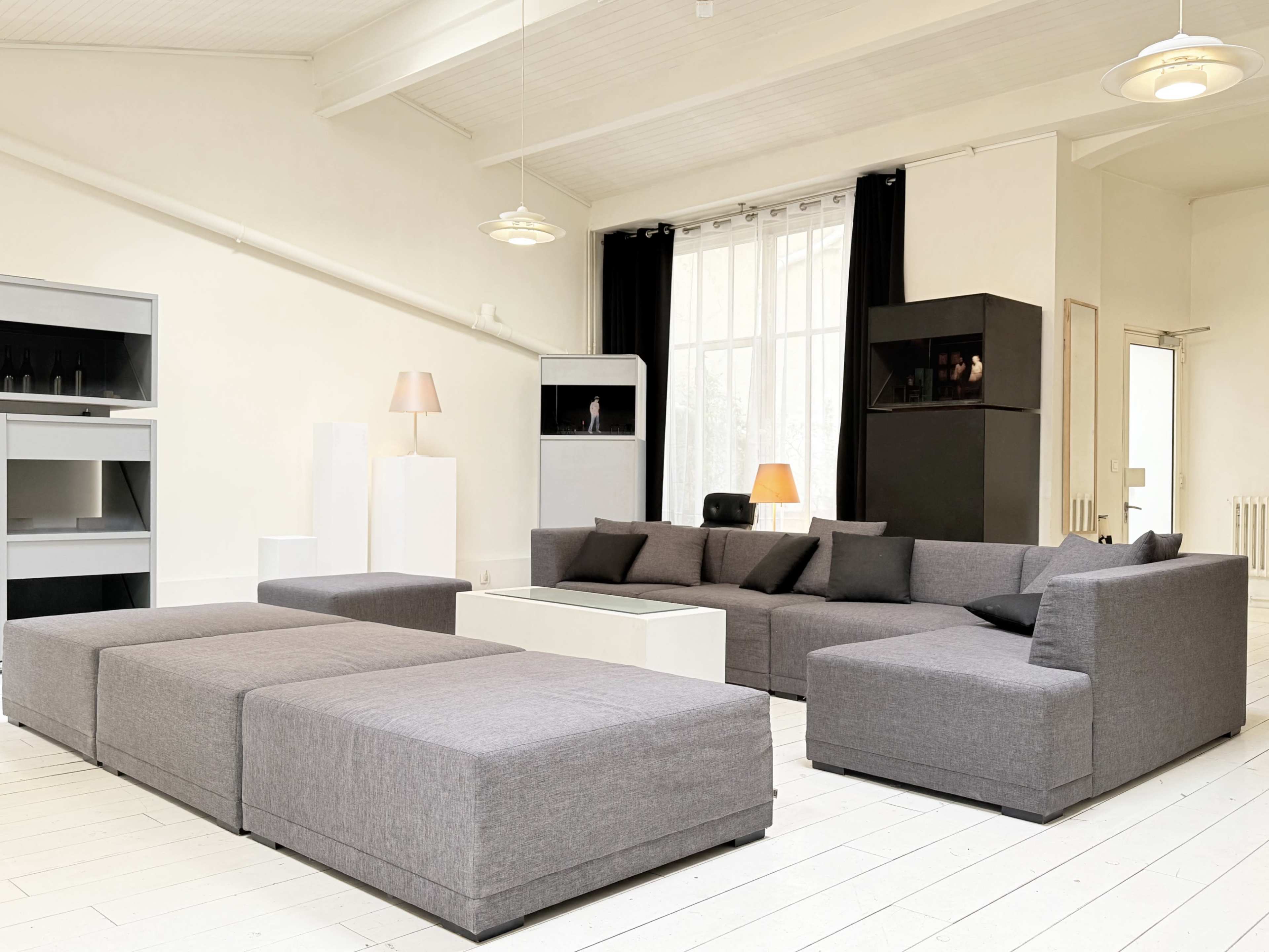 The image shows a spacious modern living room featuring a gray sectional sofa, a low coffee table, and large windows with light curtains.