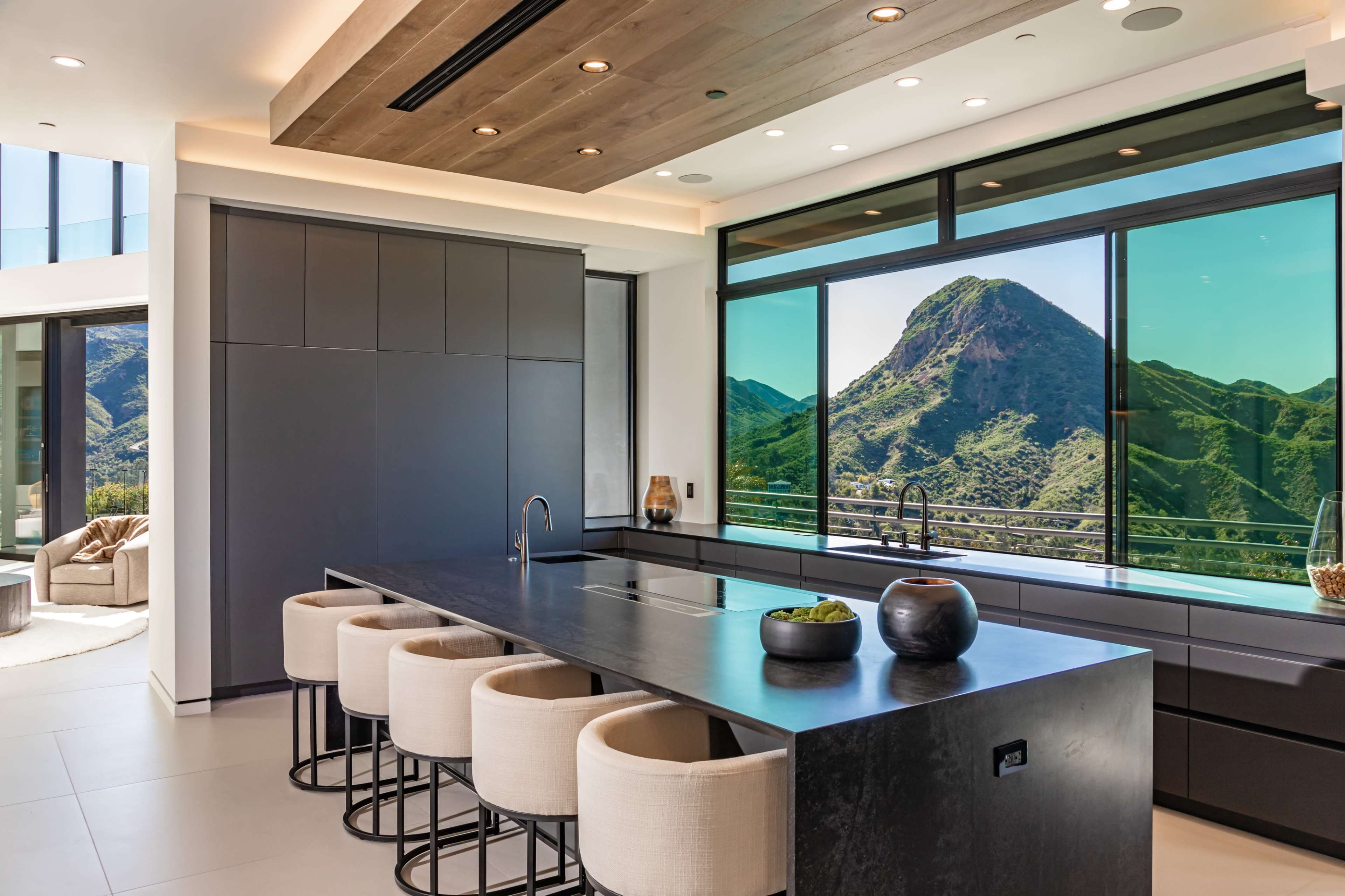 A modern kitchen features a large island with bar stools, and floor-to-ceiling windows showcase a view of a mountainous landscape.