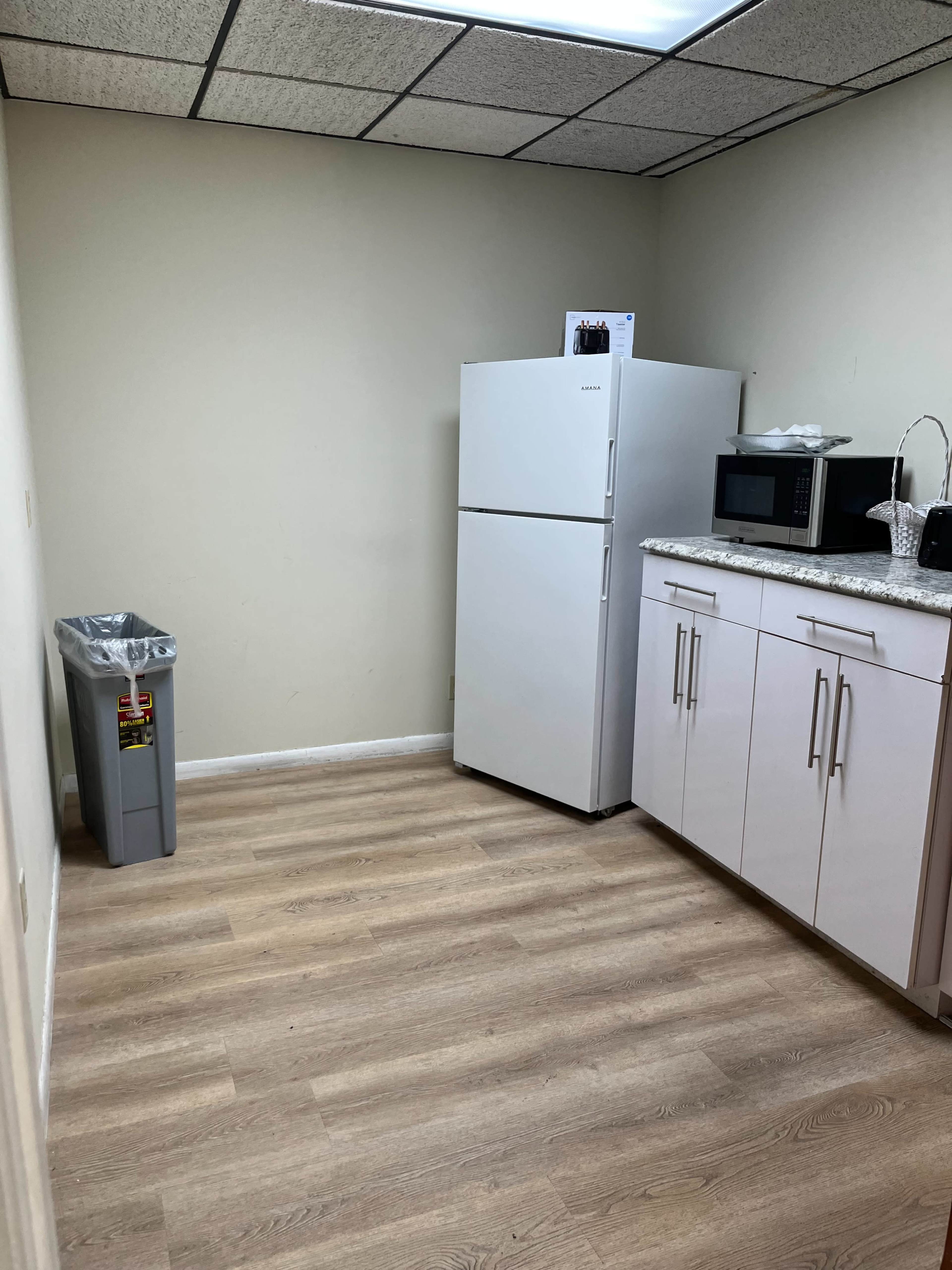 A small kitchenette with a white refrigerator, a microwave, and a gray trash can against a plain wall.
