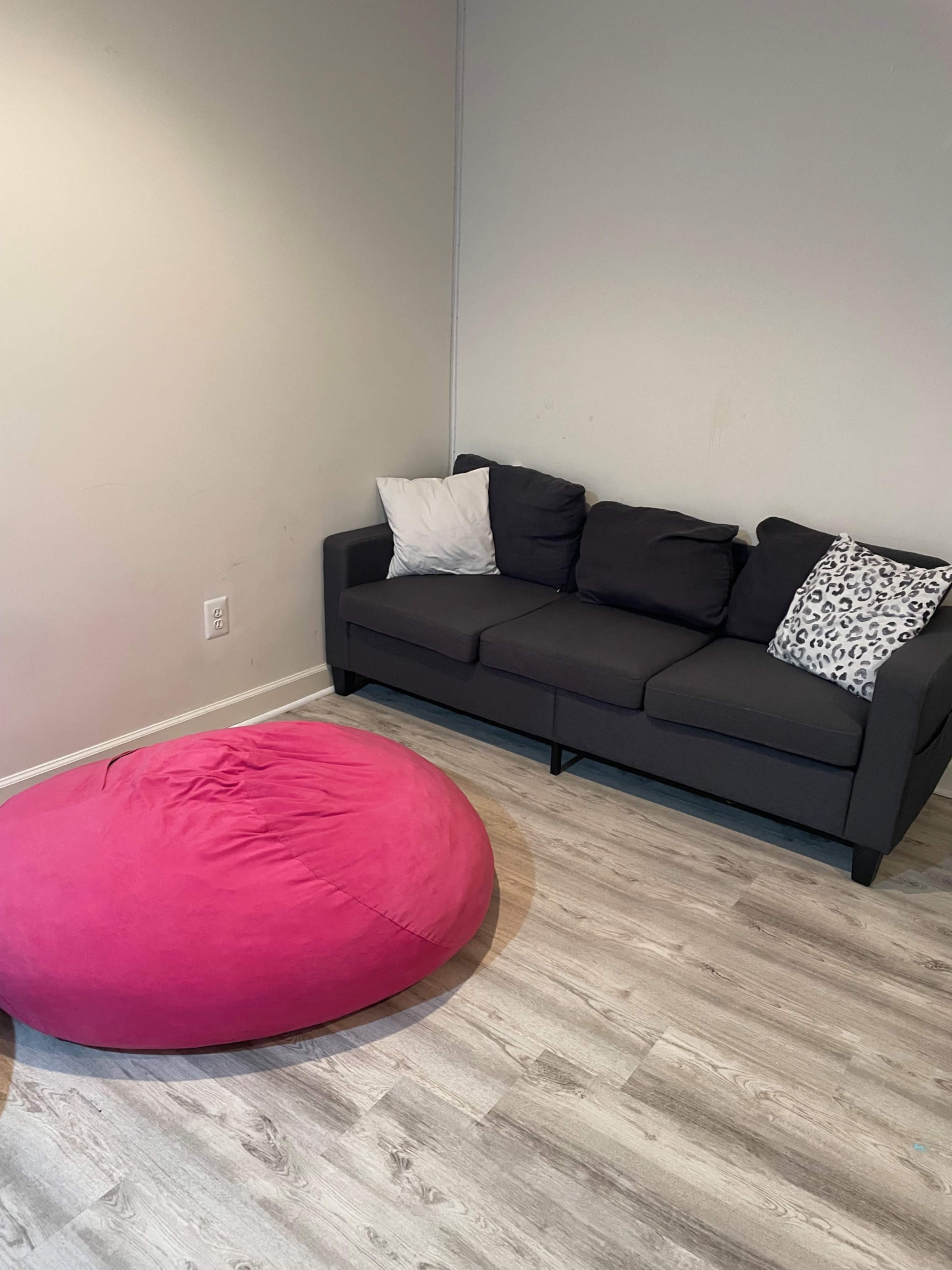 A gray sofa with black and white pillows is positioned next to a large pink bean bag chair on a wooden floor.