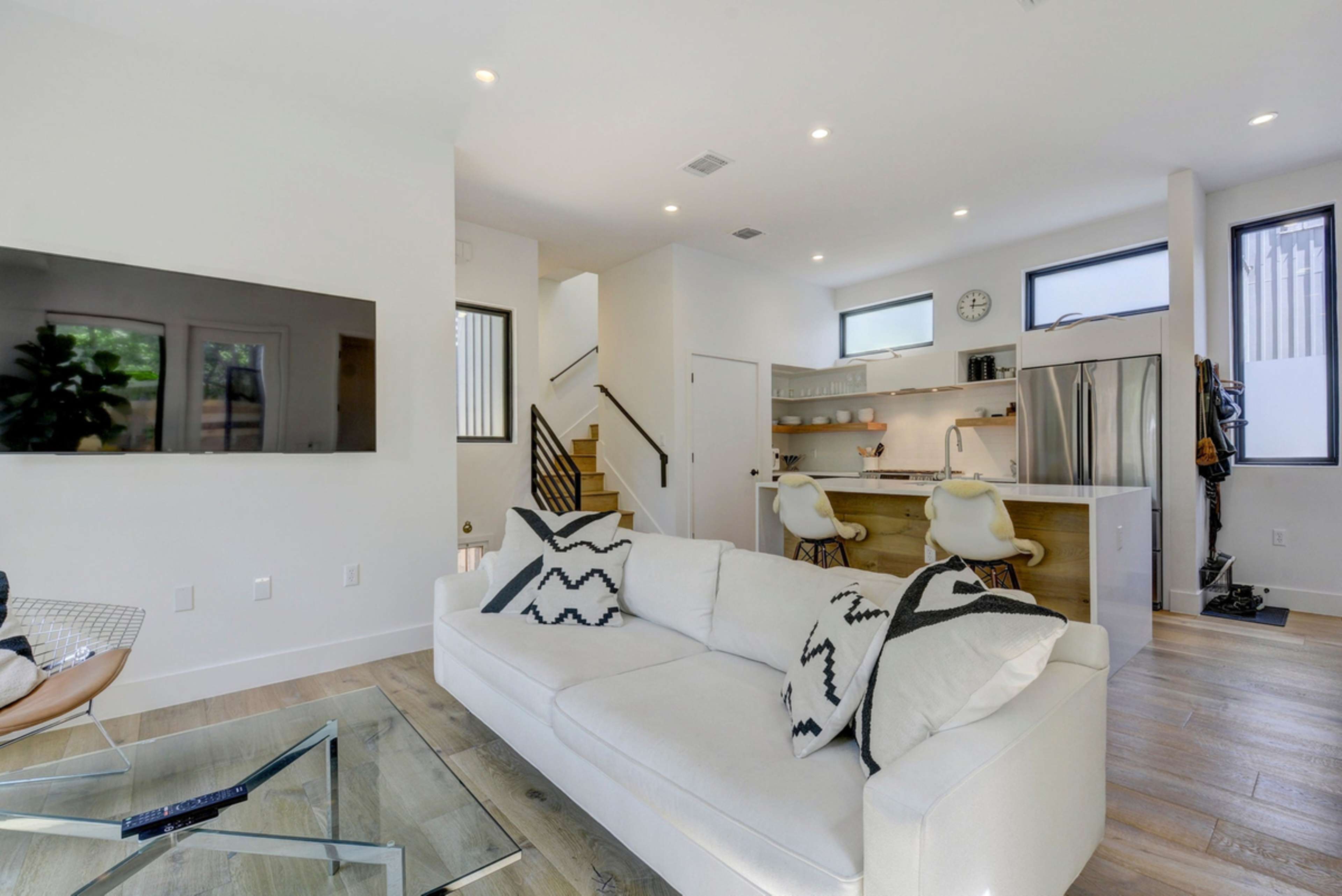 A modern living room features a white sofa with patterned throw pillows, a glass coffee table, and an open kitchen visible in the background.