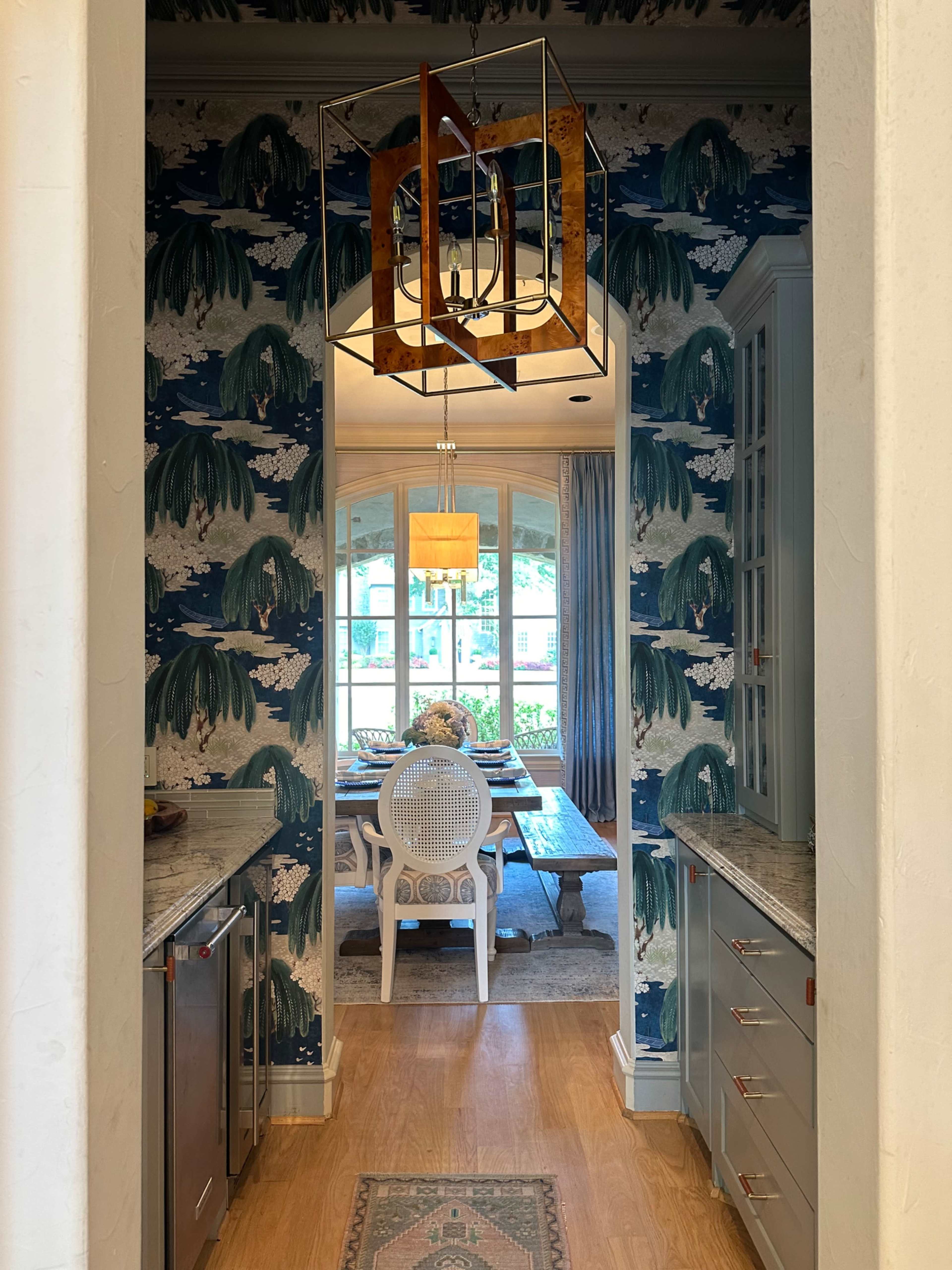 A hallway leads to a dining area with a patterned wallpaper, hardwood floor, and a large chandelier above a table.