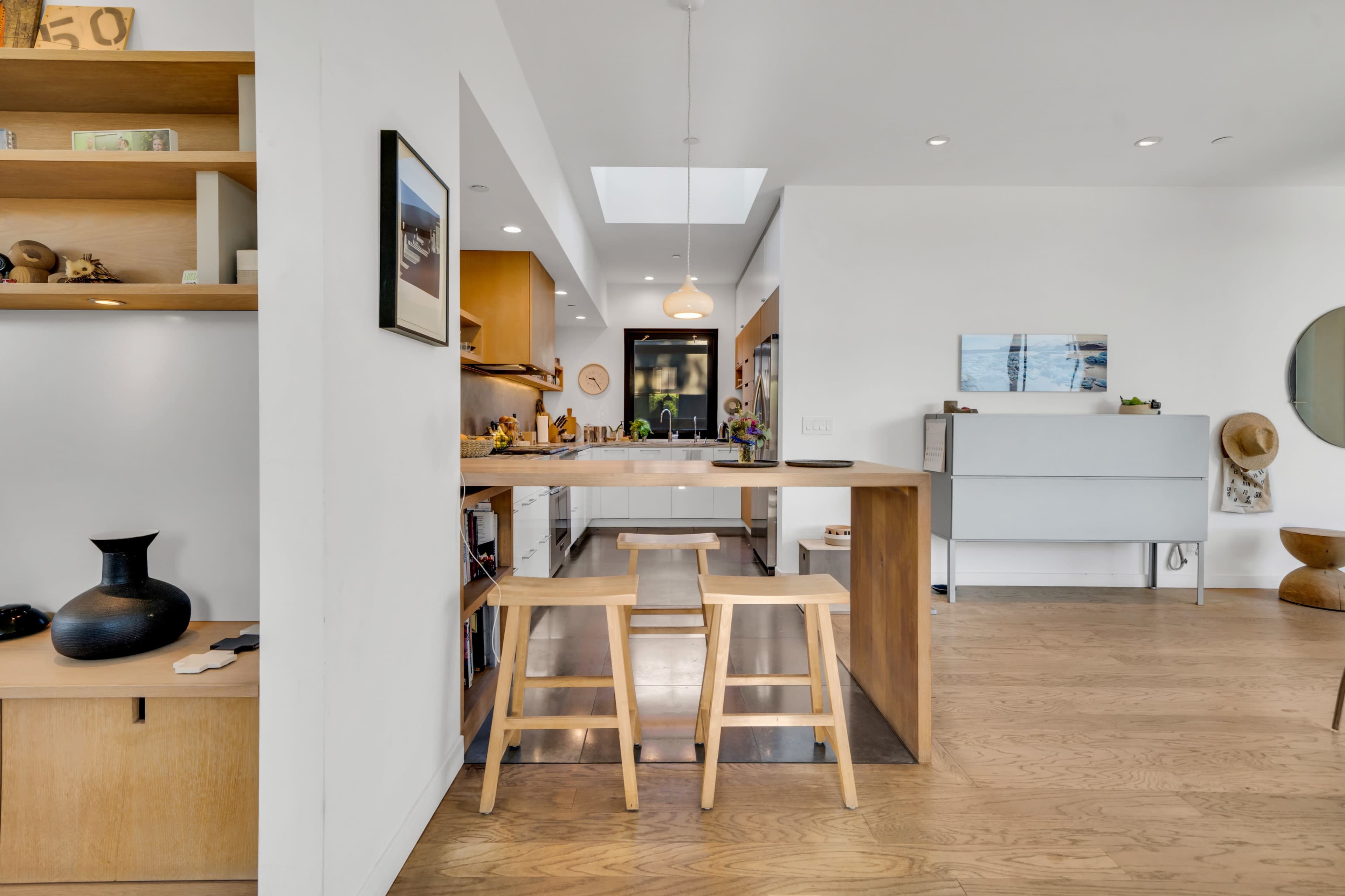 A modern kitchen with an island and two wooden stools, featuring open shelving and a bright skylight above.