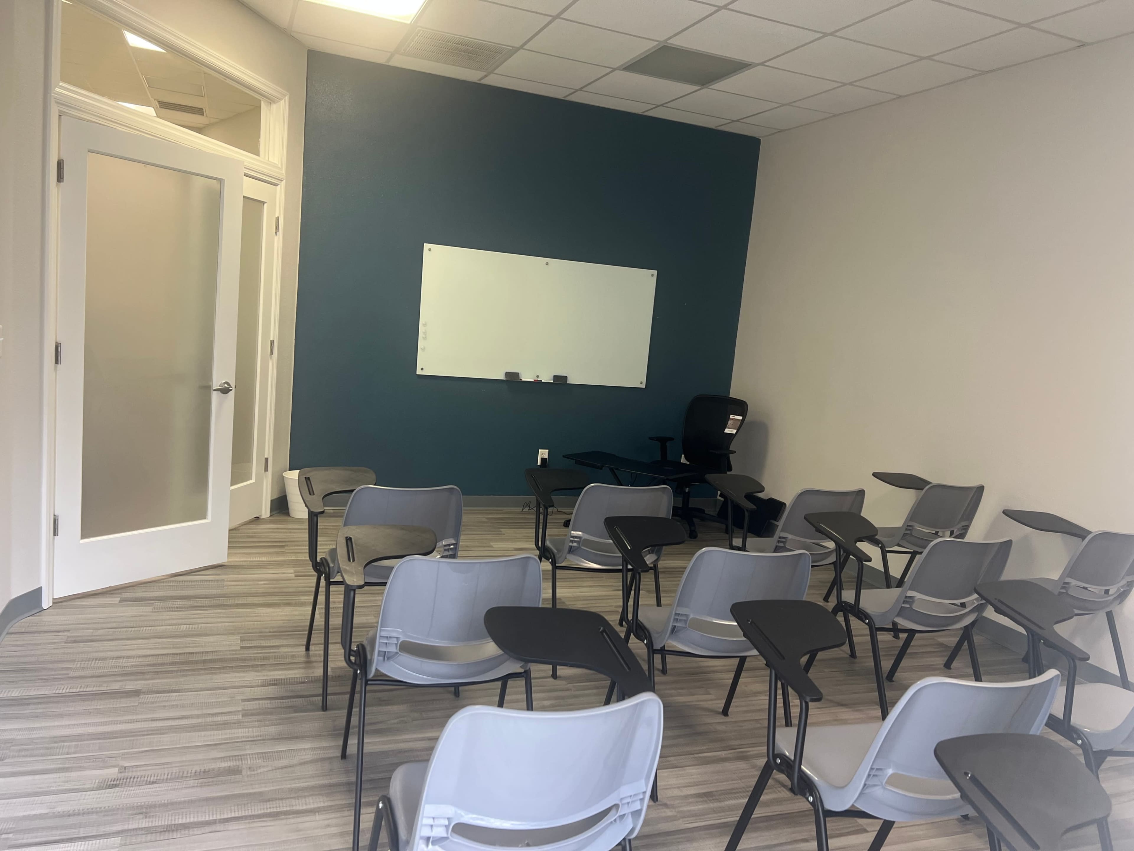 The image shows a classroom with rows of chairs and a whiteboard on a teal wall.