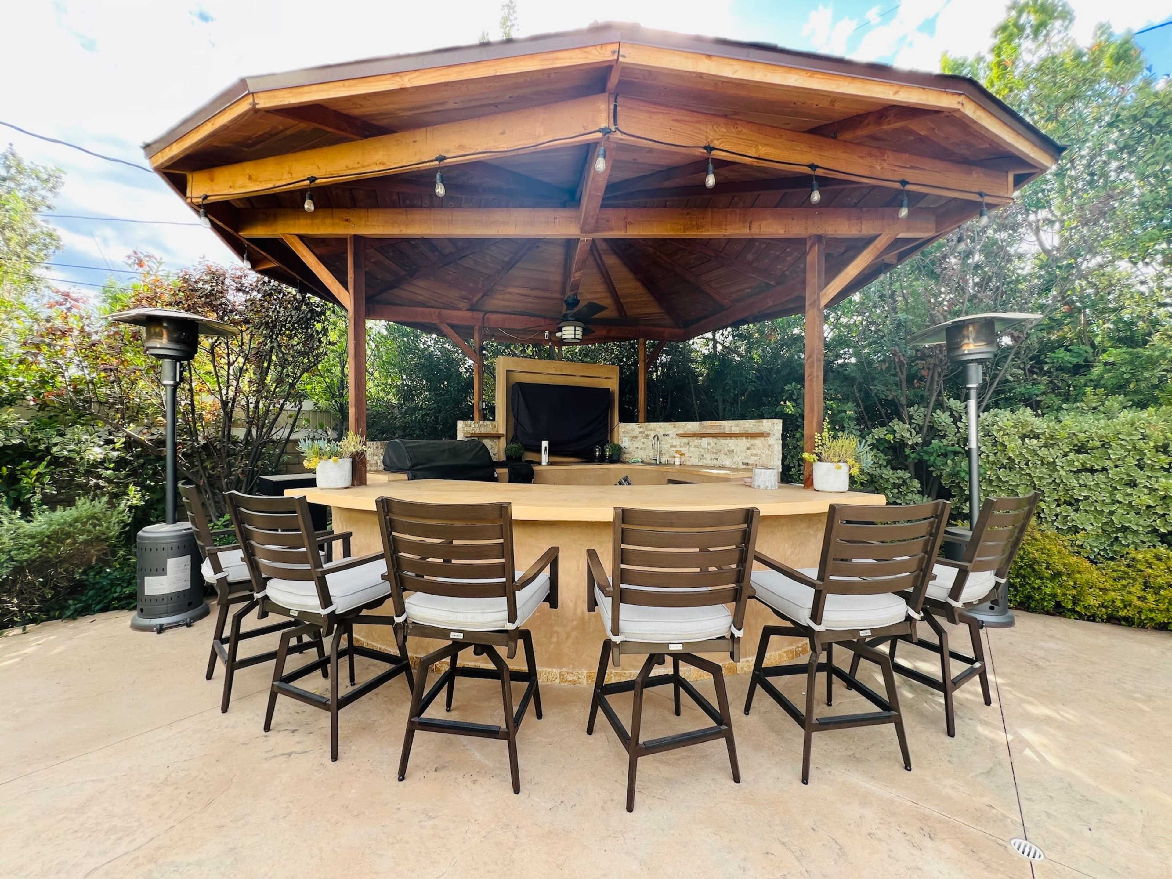 A wooden gazebo with a central table surrounded by eight tall-backed chairs and outdoor heaters, set in a landscaped area with greenery.