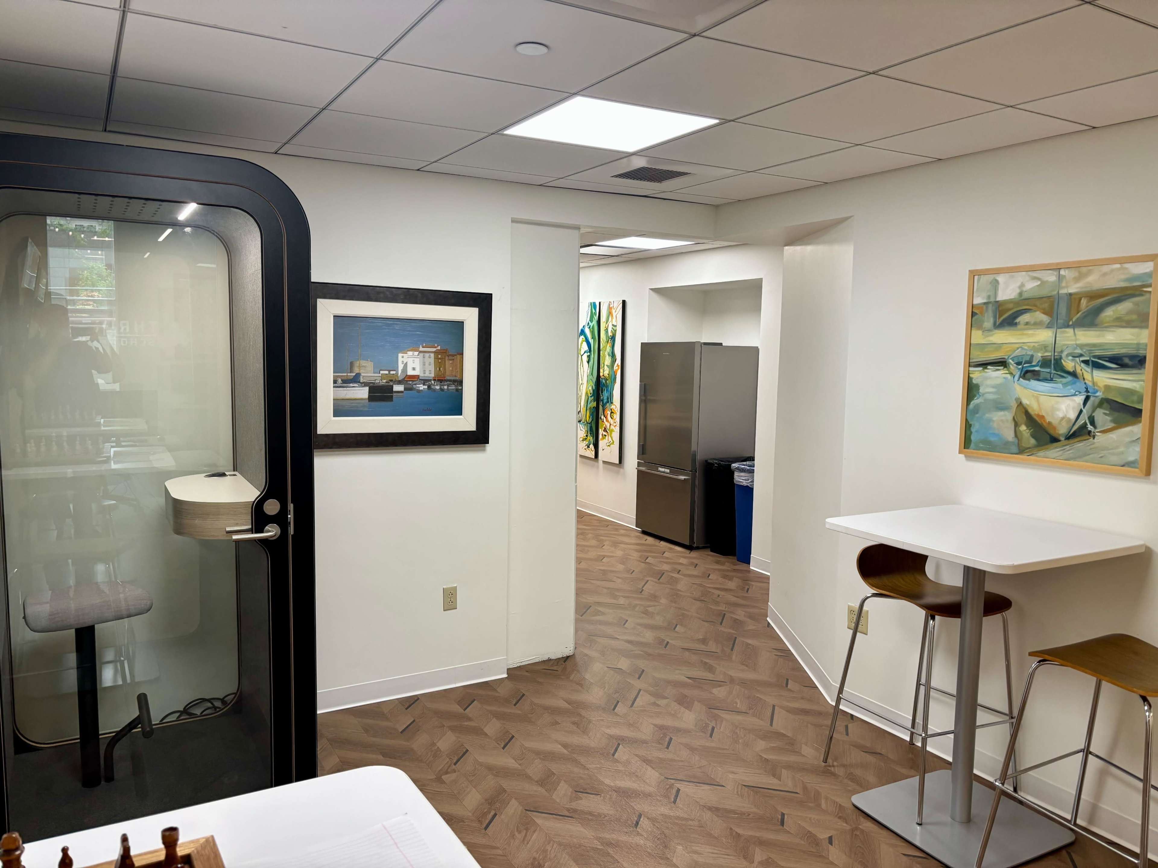 The image shows a modern office space featuring a glass phone booth, artwork on the walls, a kitchenette with a refrigerator, and a small dining area with stools and tables.