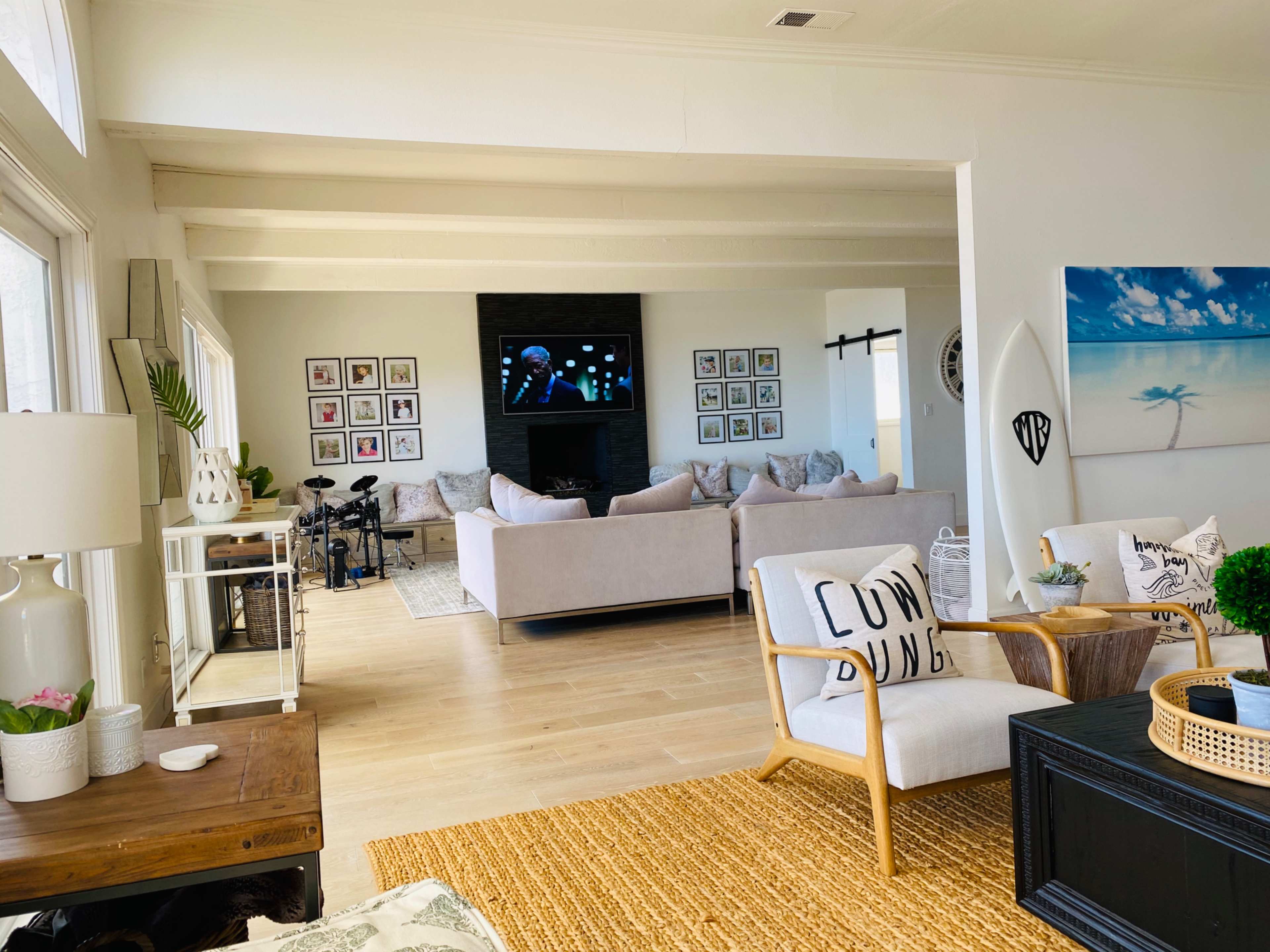 The image shows a bright living room with a large TV mounted on a wall, surrounded by framed pictures and a comfortable seating area featuring a light-colored sofa and wooden furniture.