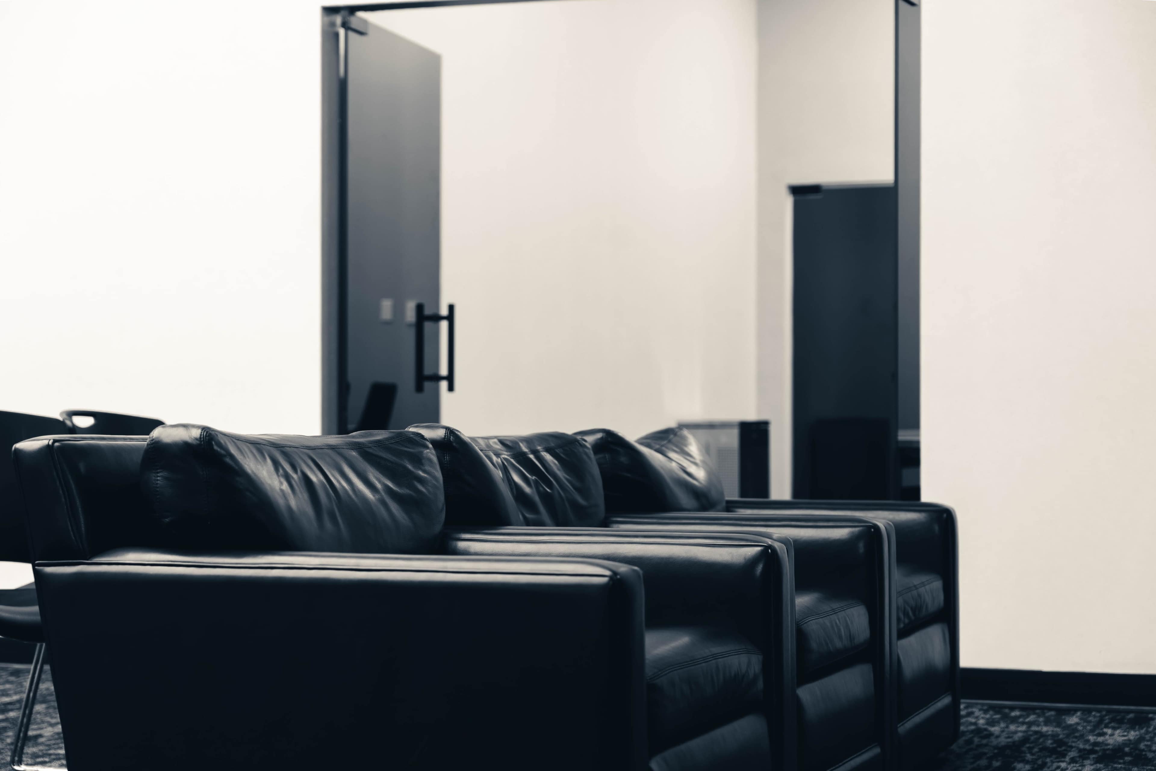 A row of black leather chairs is positioned in a minimalist room, with an open doorway and a mirrored wall in the background.