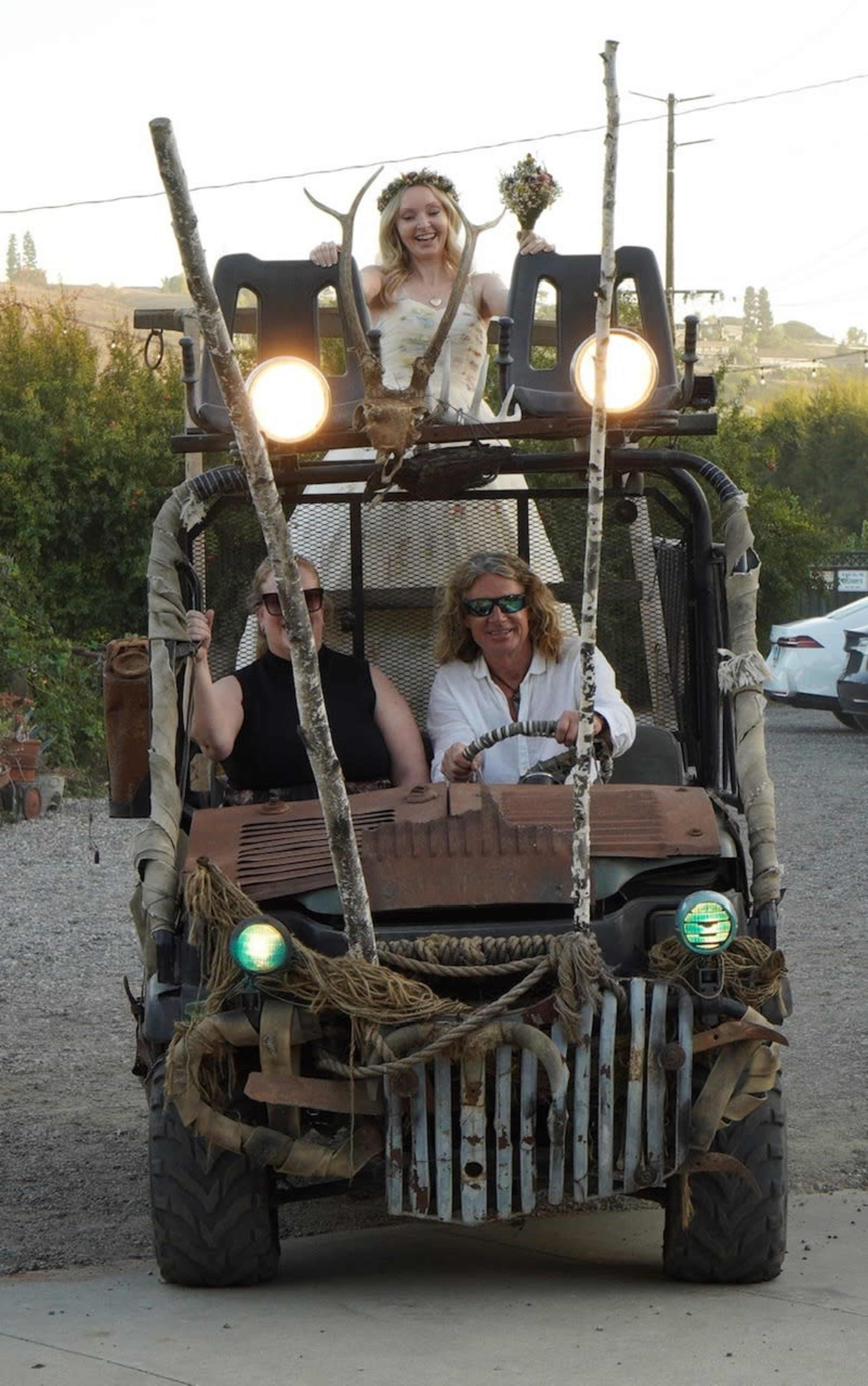 A group of three people is riding in a modified ATV decorated with tree branches and lights, while one person stands on top holding a bouquet.