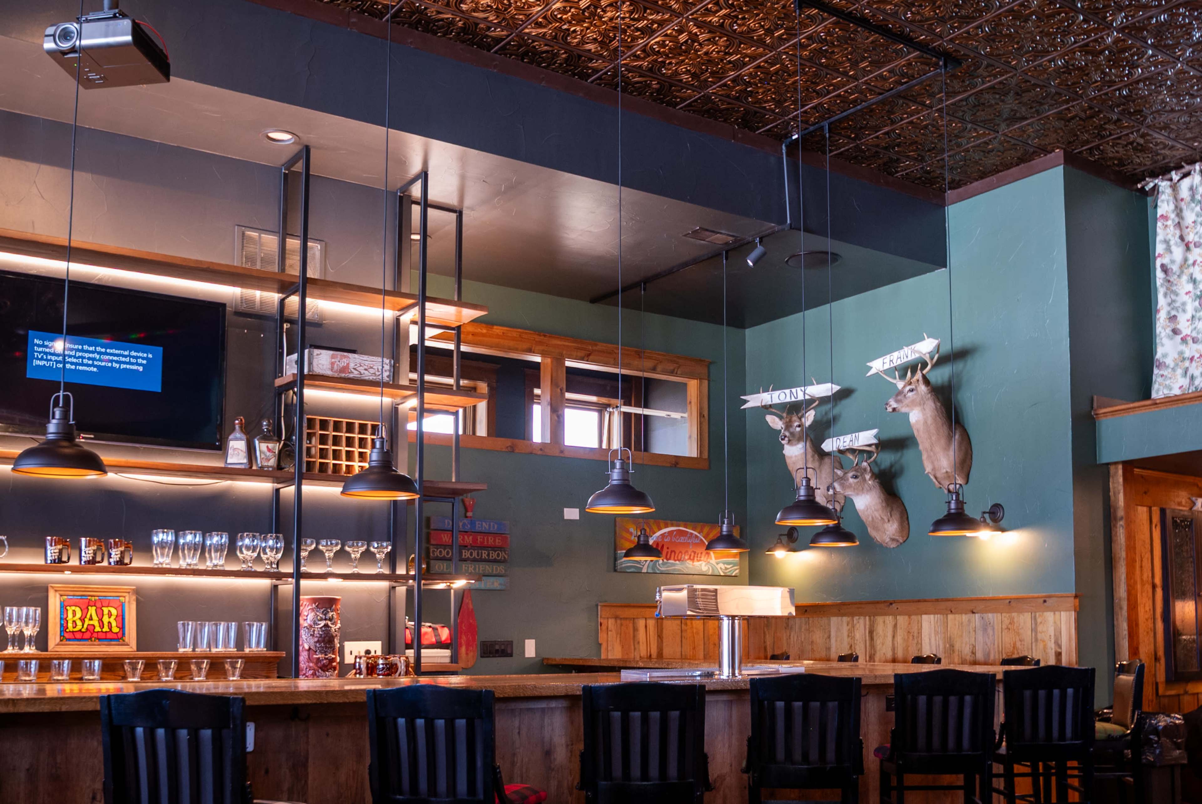 The interior of a bar features wooden furnishings, mounted deer heads on the wall, and a well-stocked counter with various glassware.