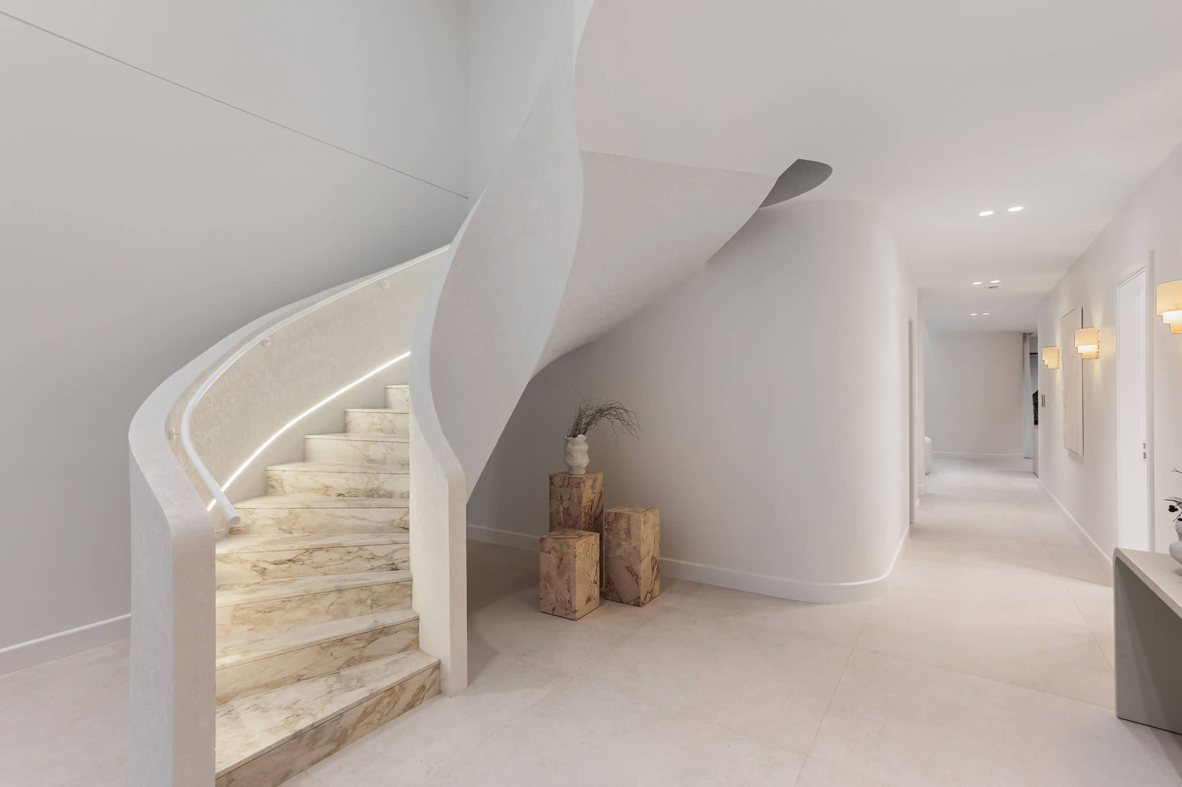 The image depicts a modern entryway with a curved staircase made of marble and minimalist decor, featuring light-colored walls and a series of recessed lights.