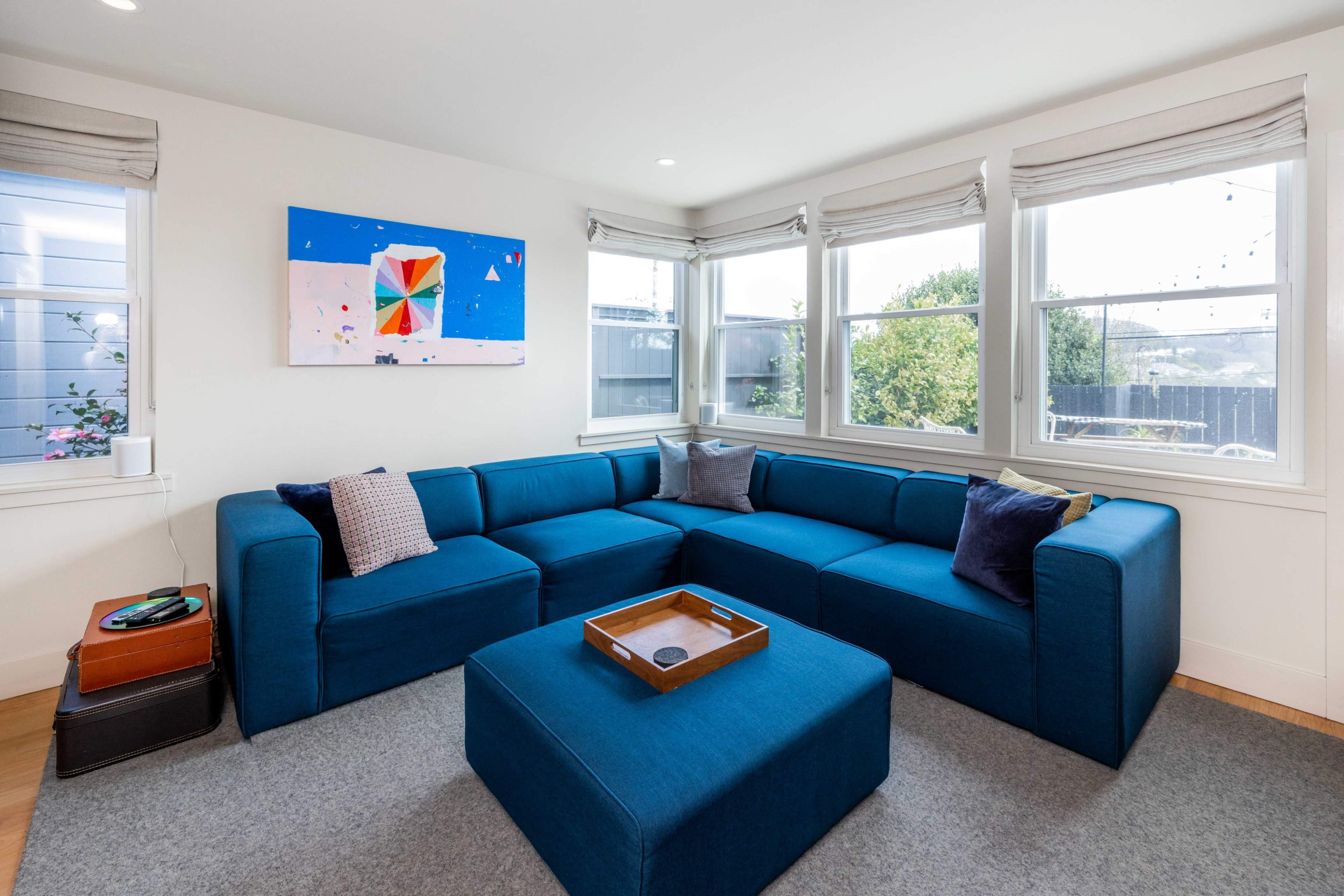A modern living room with a large blue sectional sofa, an ottoman, and a colorful abstract painting on the wall, illuminated by natural light from several windows.
