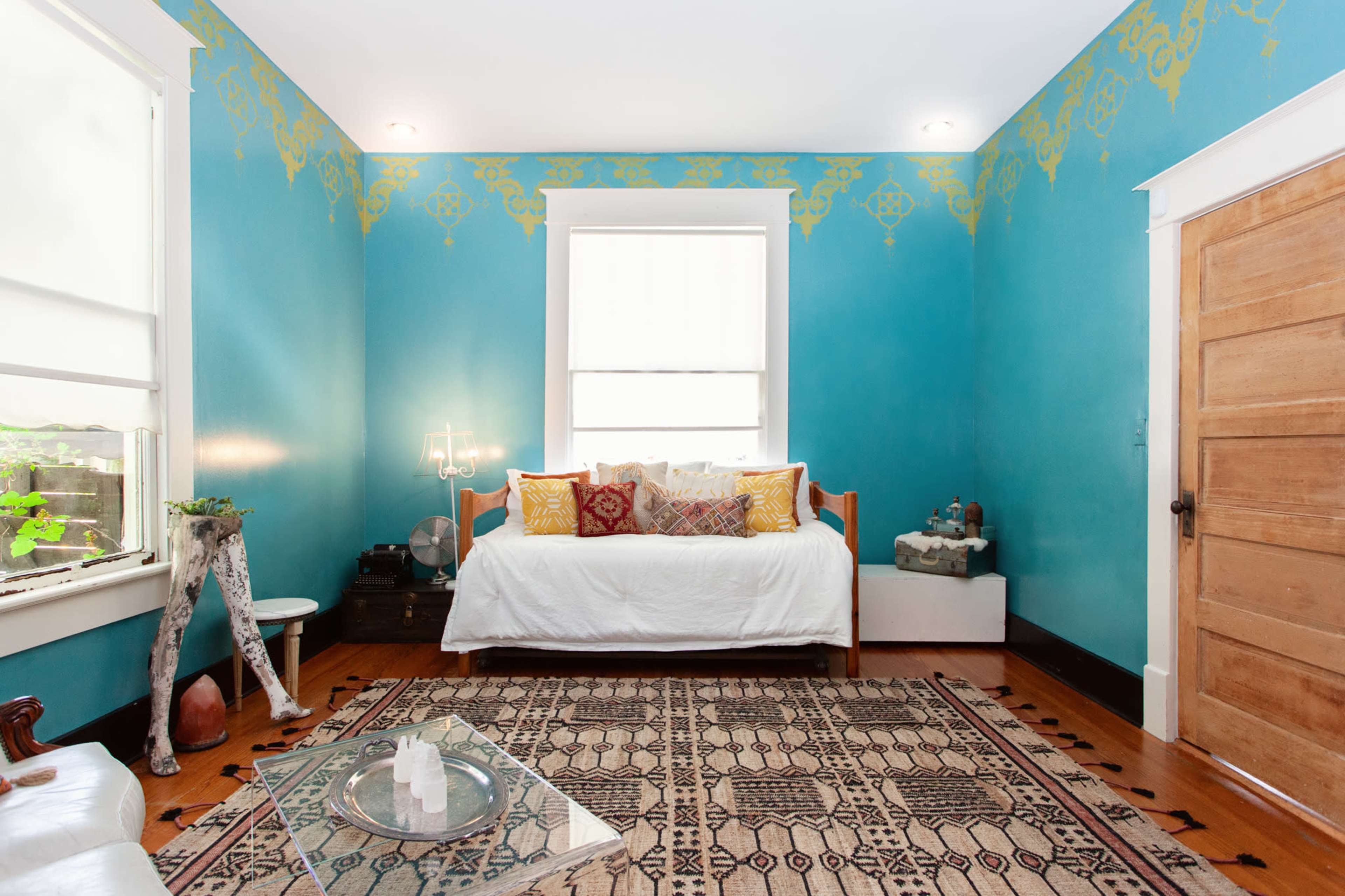 A brightly colored room features a daybed adorned with decorative pillows, a glass coffee table, and an intricately patterned rug on polished wooden floors.