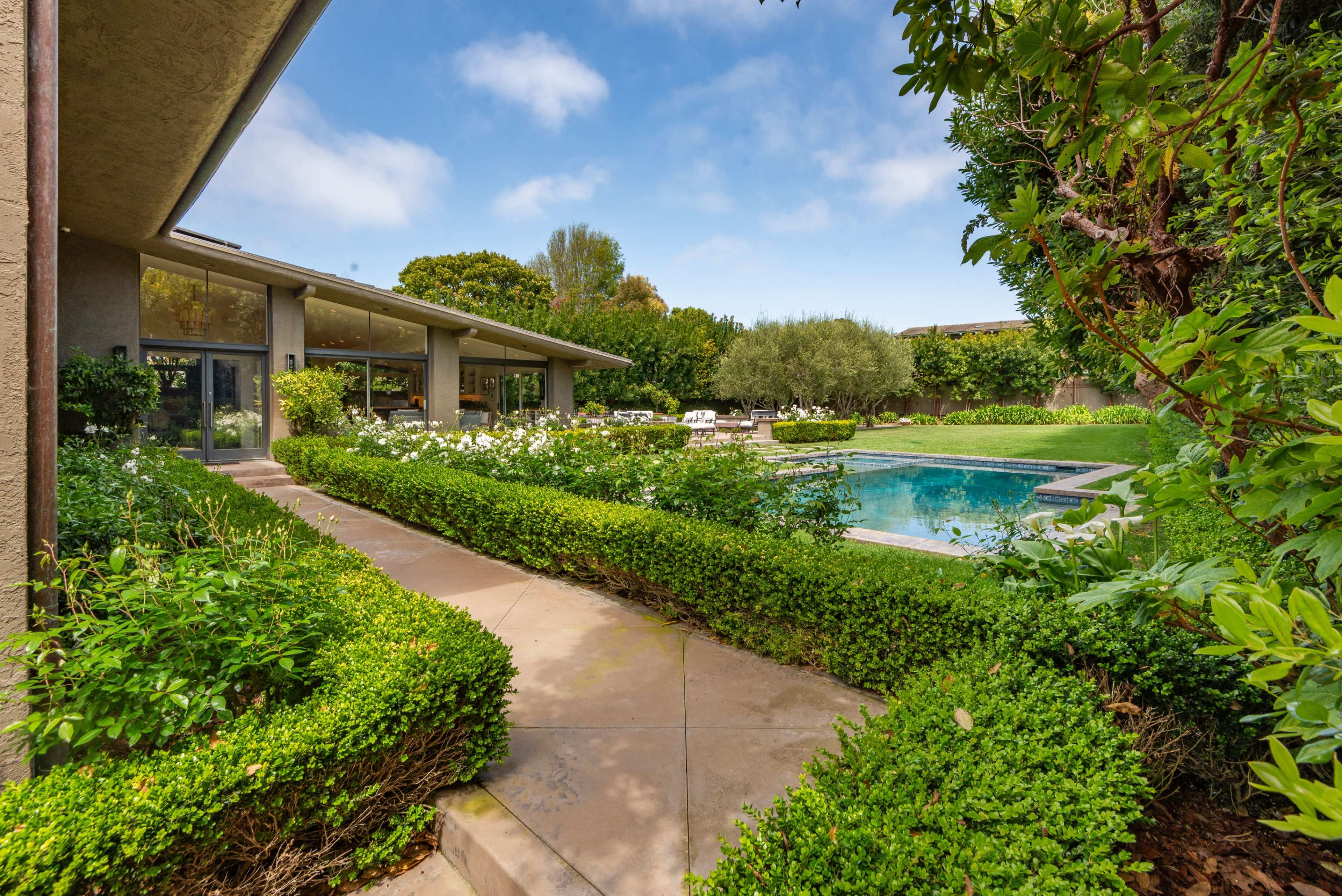 A landscaped pathway leads to a backyard with a swimming pool, surrounded by greenery and flowering plants.