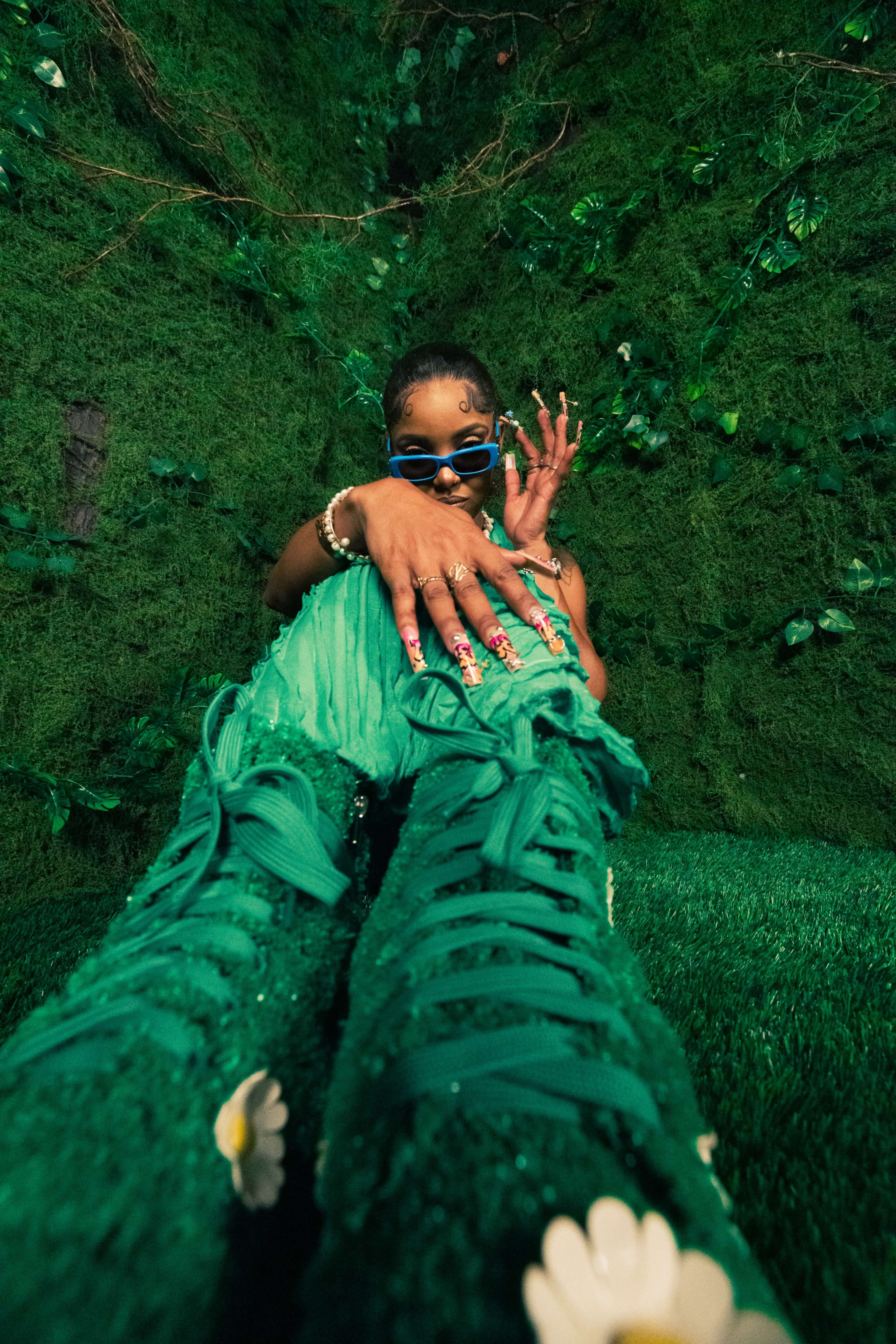 A person wearing green textured clothing sits against a backdrop of greenery, displaying an elaborate manicure and stylish sunglasses.