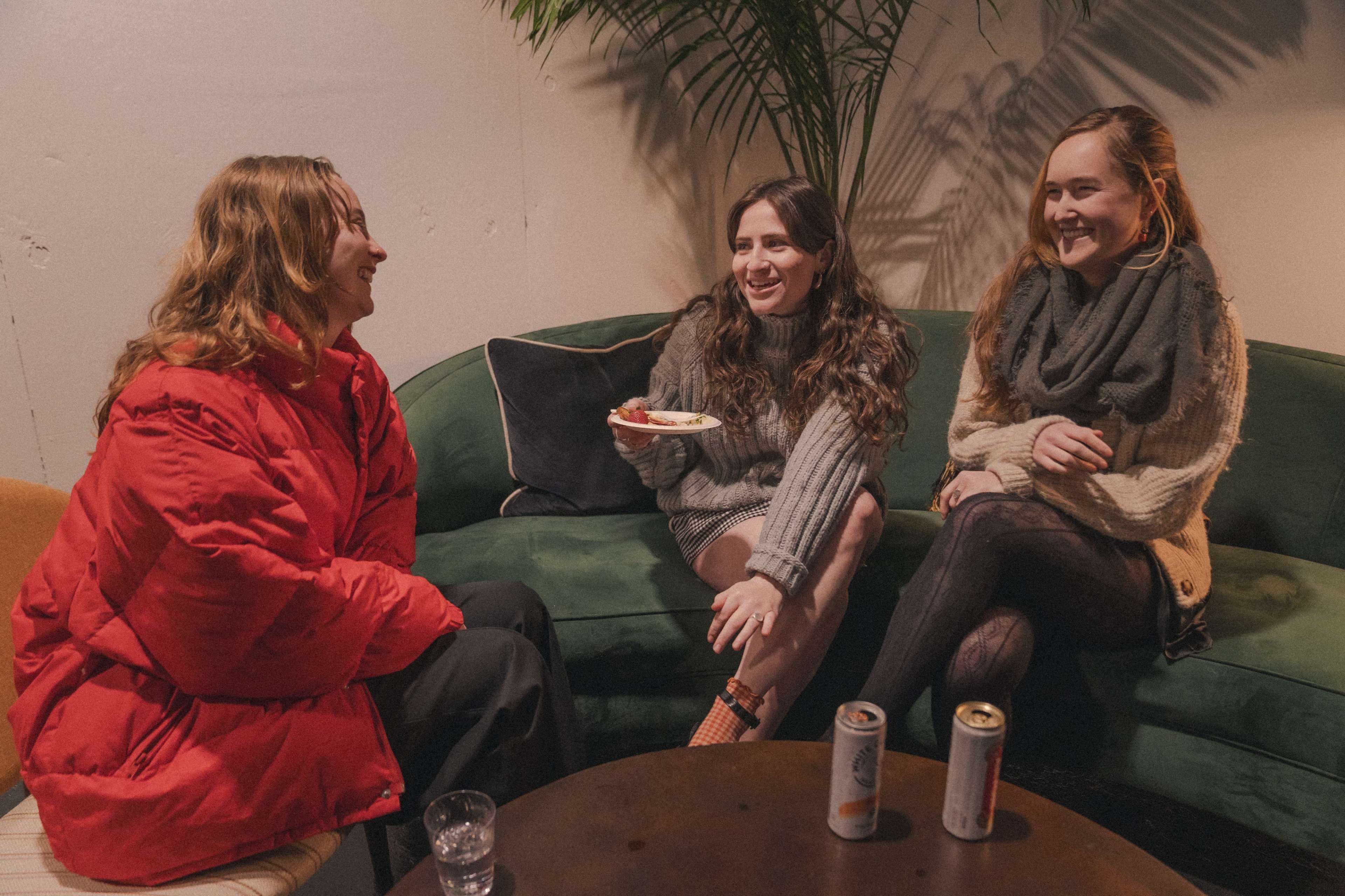 Three women are sitting on a green couch, engaged in conversation while sharing snacks and drinks.