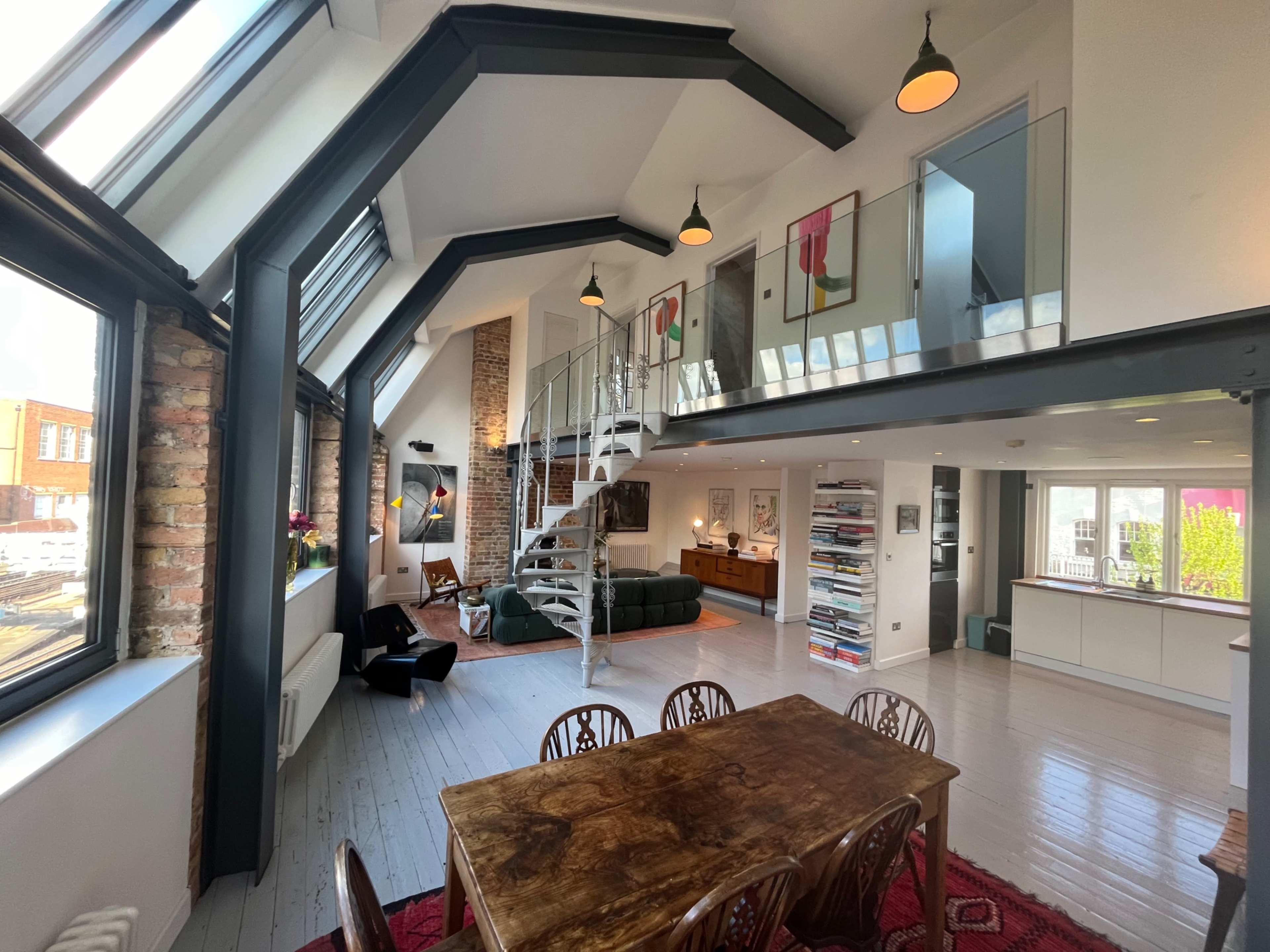The image shows a spacious, modern loft featuring a dining area with a wooden table, a living area with a green sofa, and an upper level accessible by a spiral staircase.