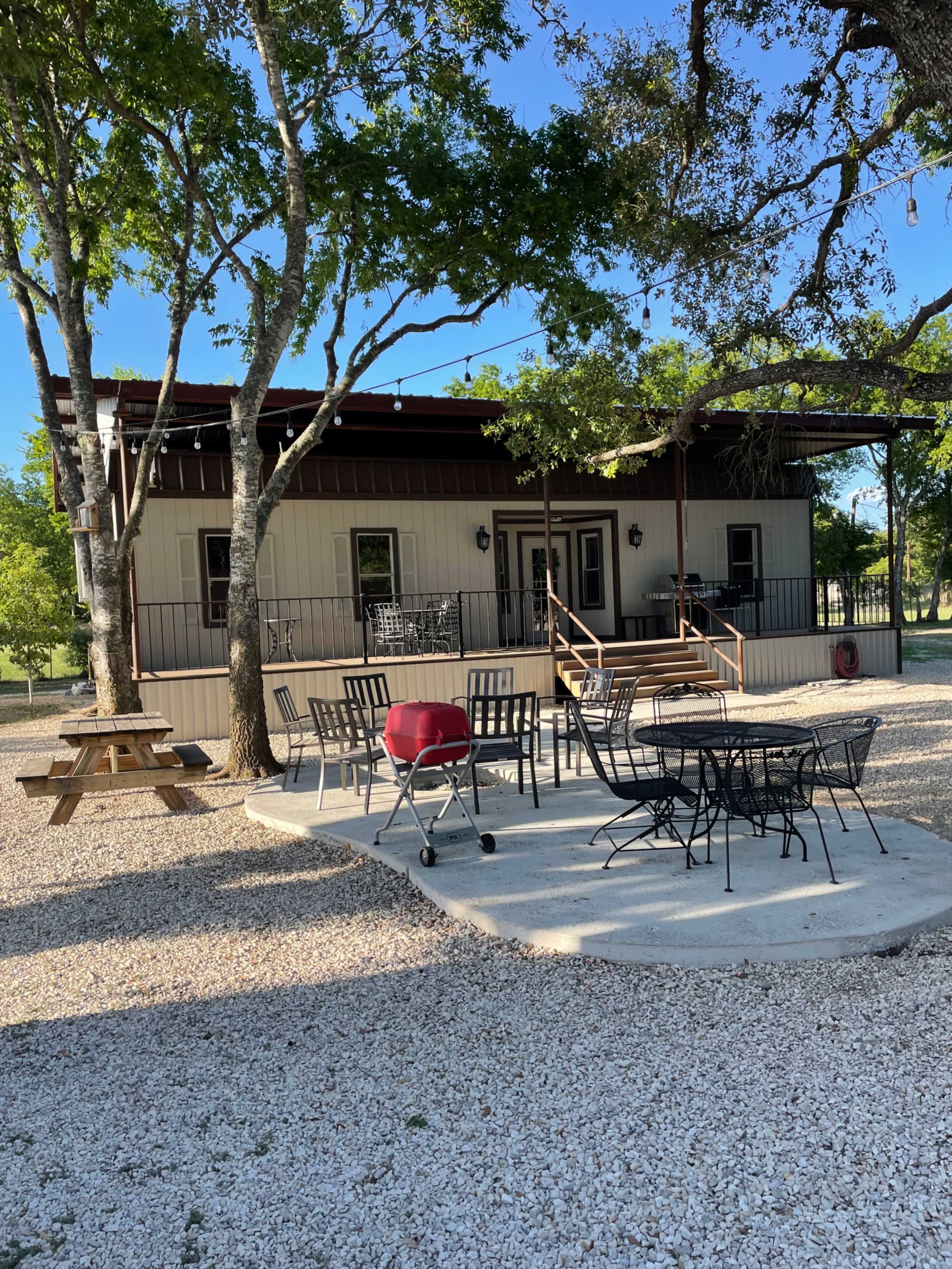 A modern building with a covered porch is surrounded by outdoor seating and a grill, set in a gravel area with trees nearby.