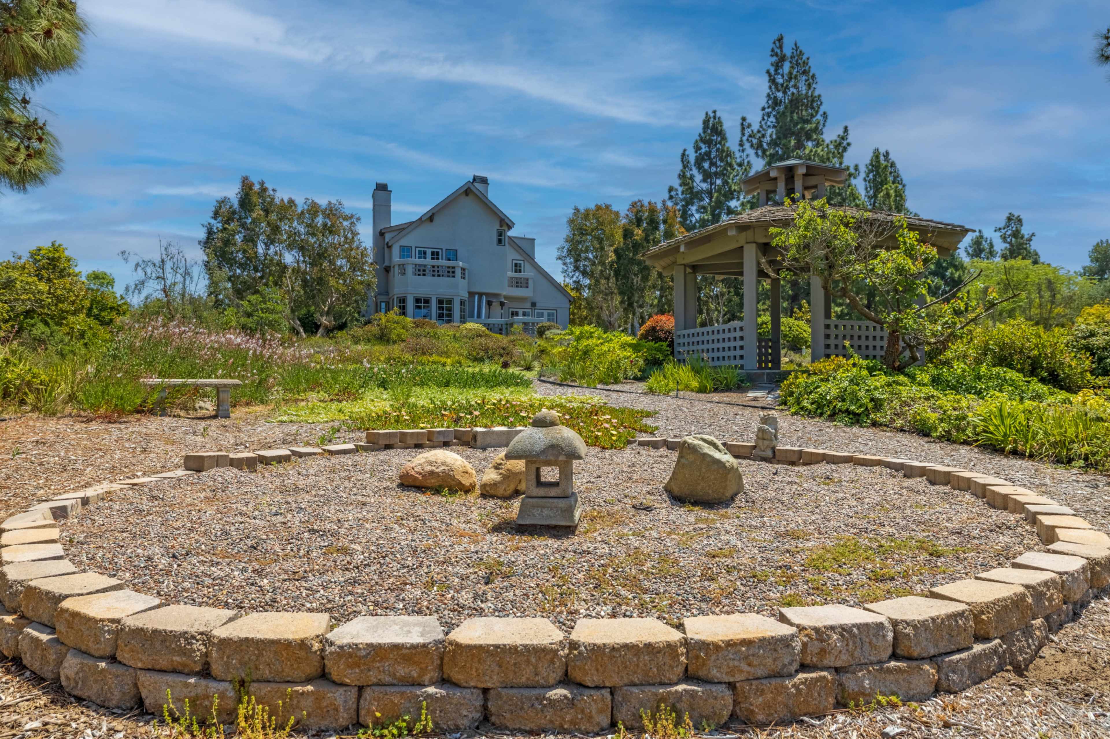 A landscaped garden features a circular stone arrangement with rocks and a lantern, surrounded by vibrant vegetation and a large house in the background.