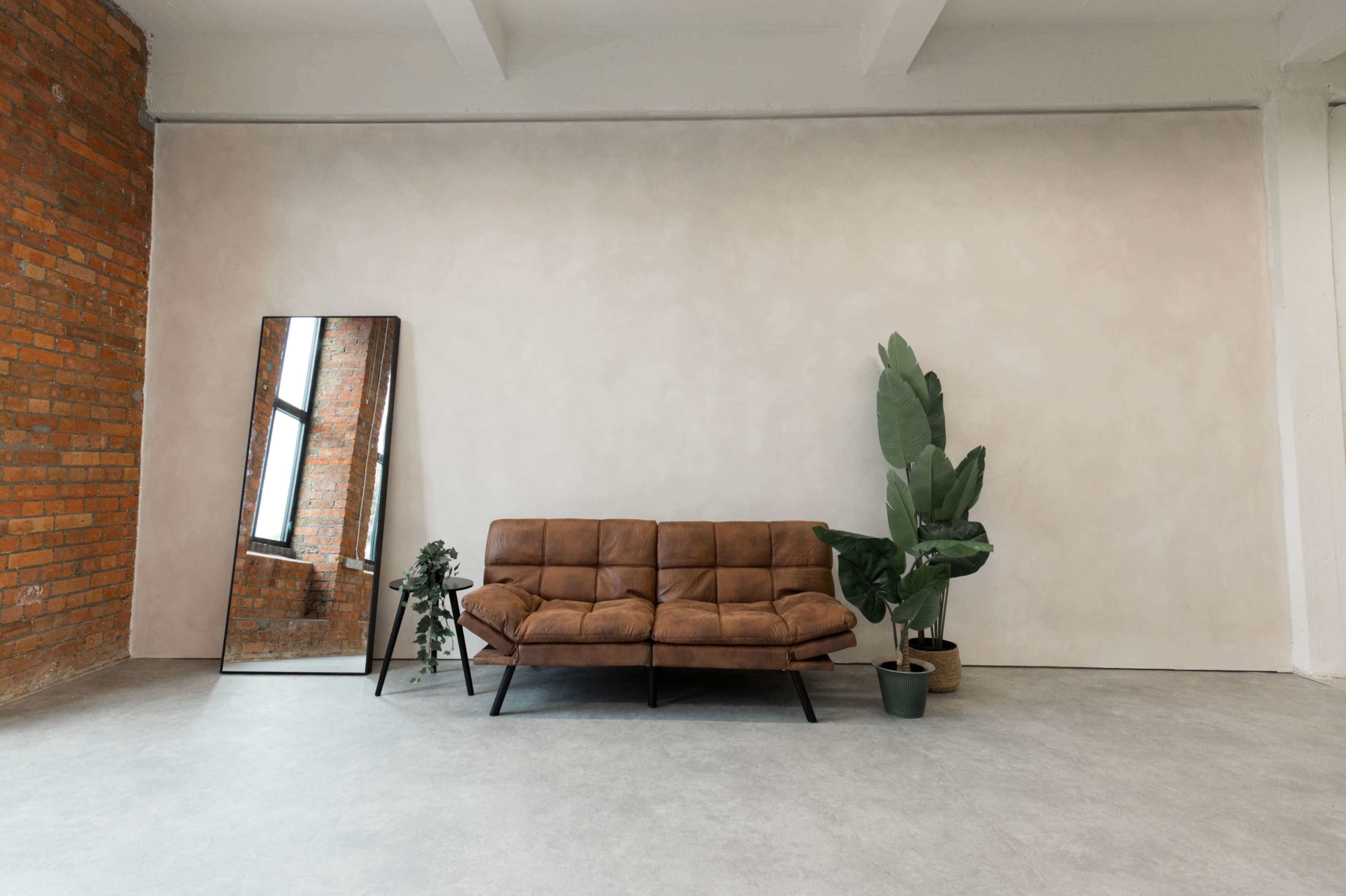 A brown leather sofa is positioned against a light-colored wall, accompanied by a mirror, a potted plant, and a small side table in a spacious room with brick accents.