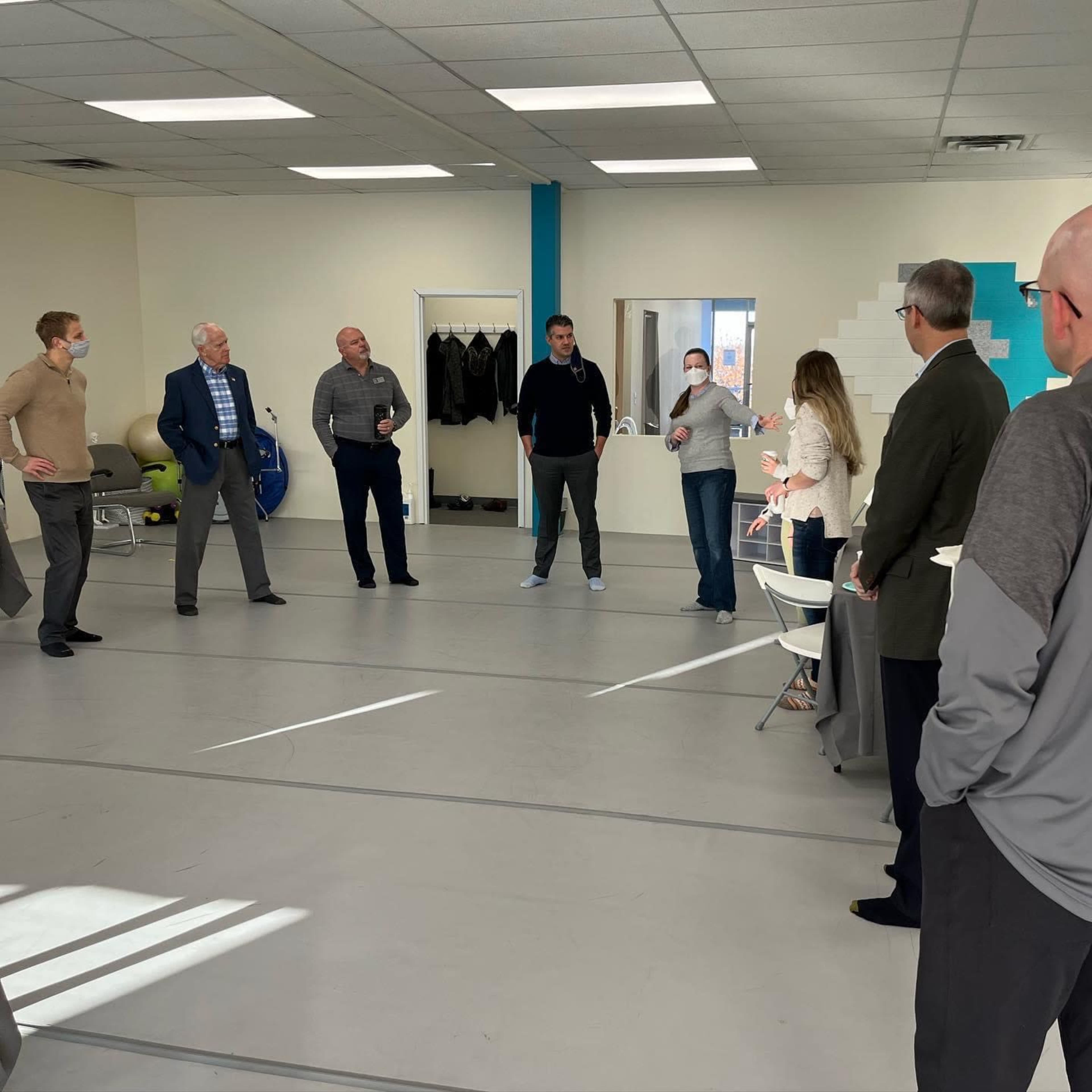 A group of eight people stands in a semi-circle in a spacious room, engaged in a discussion while wearing masks.