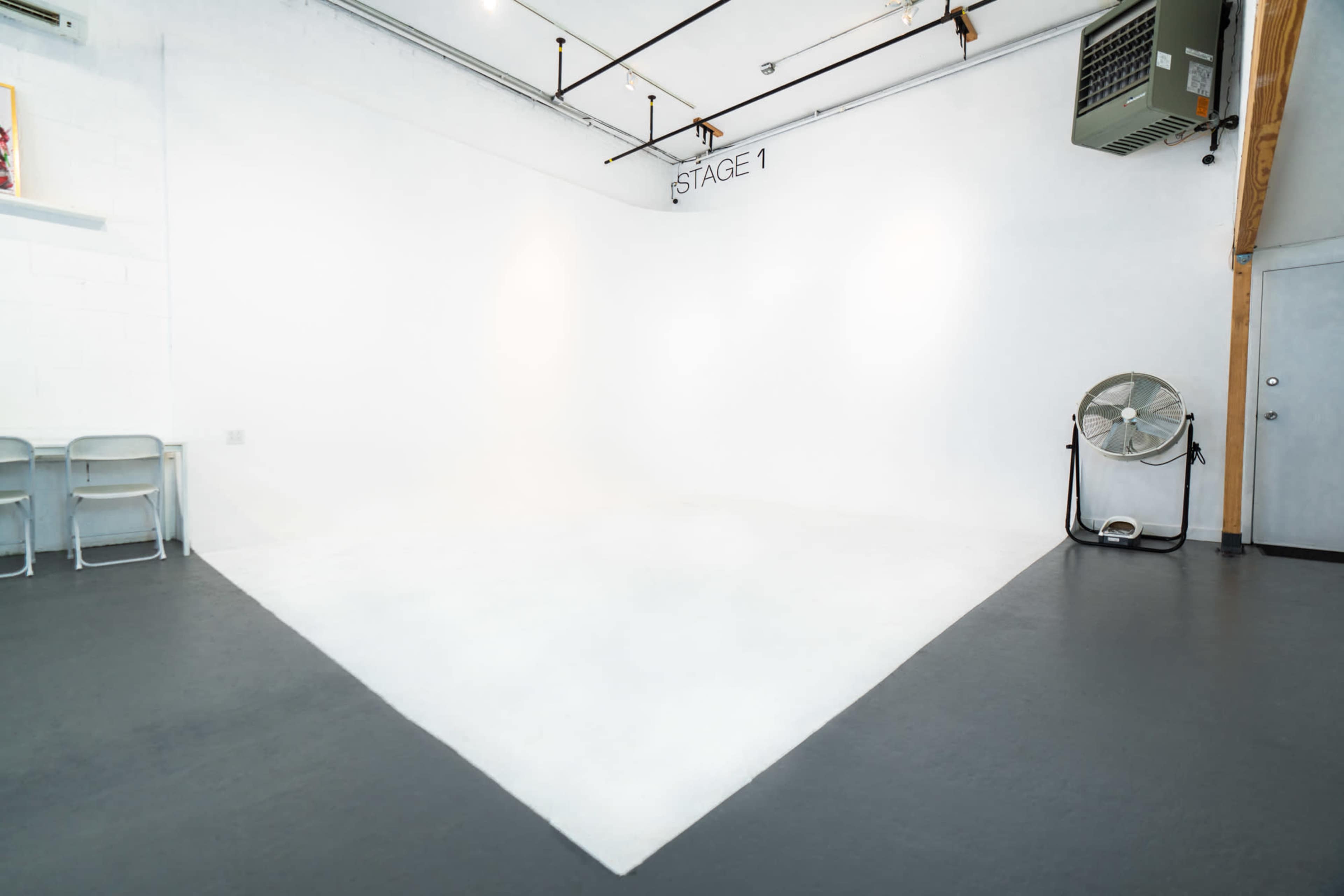 A minimalist studio space with a large white backdrop labeled "STAGE 1," a fan, and a door in the background.