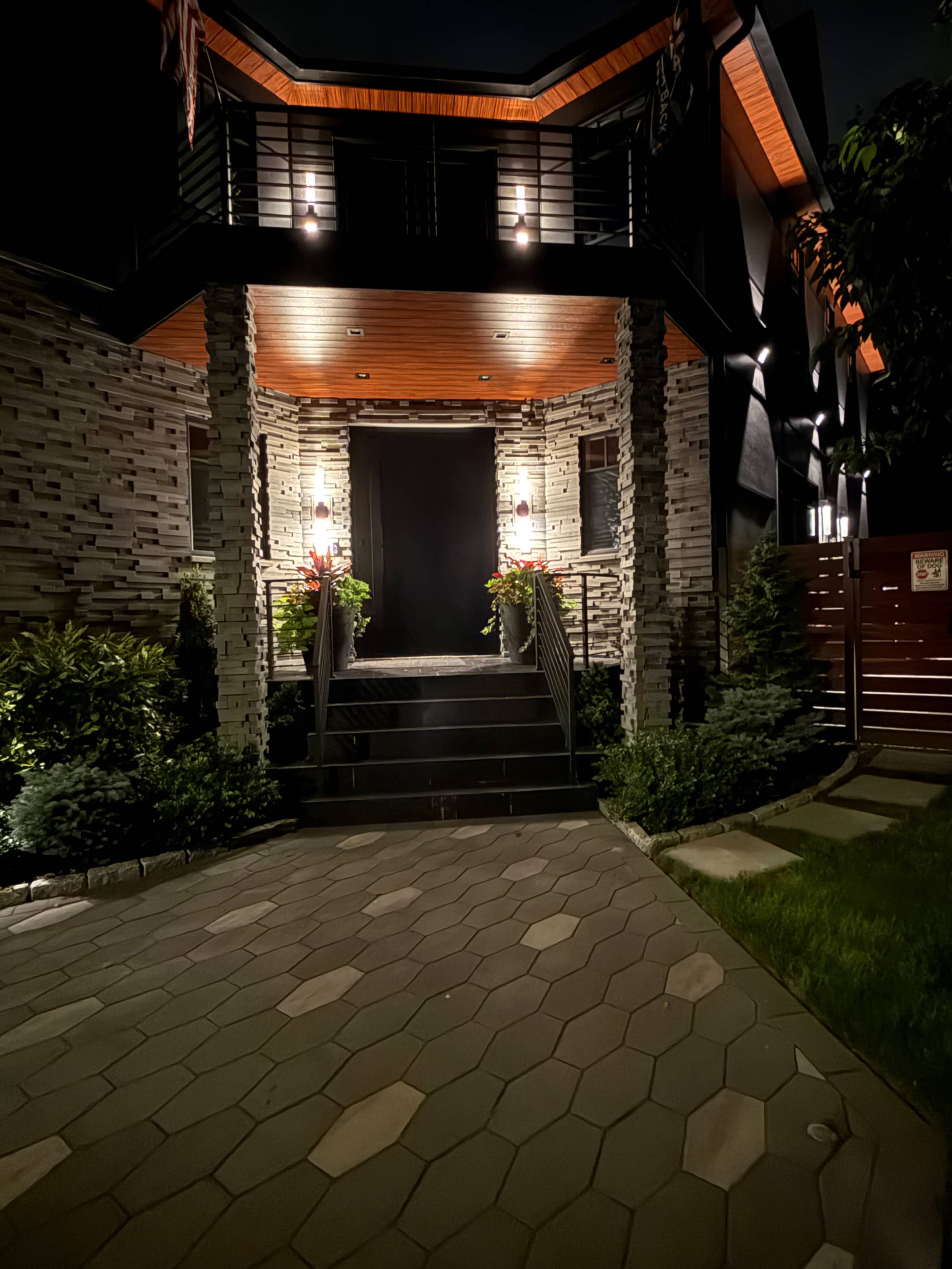 The image shows a modern entrance to a house with stone pillars, illuminated by outdoor lights, flanked by potted plants and a pathway made of hexagonal tiles.
